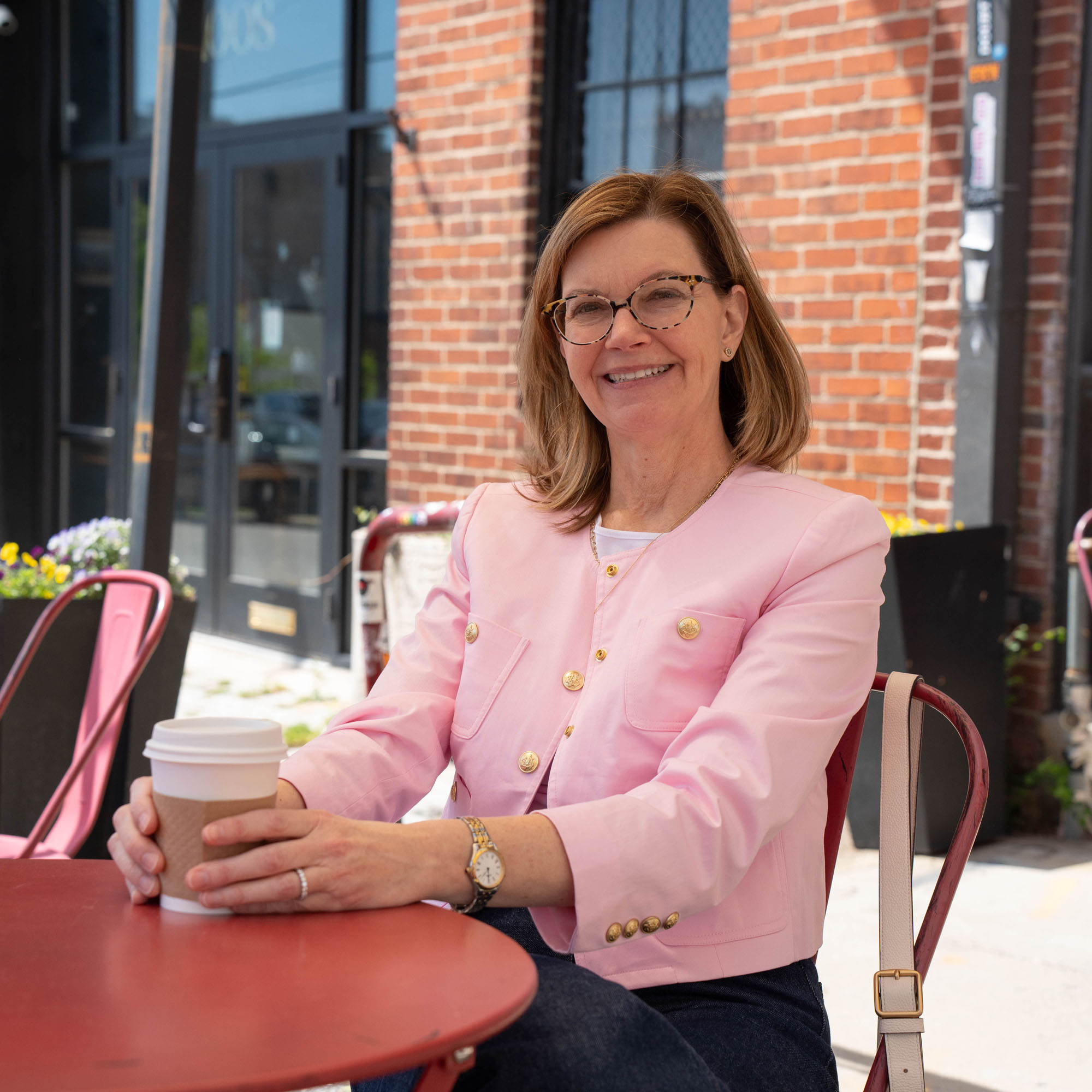 A professor poses, smiling, while seated at a red cafe table and holding a hot drink.