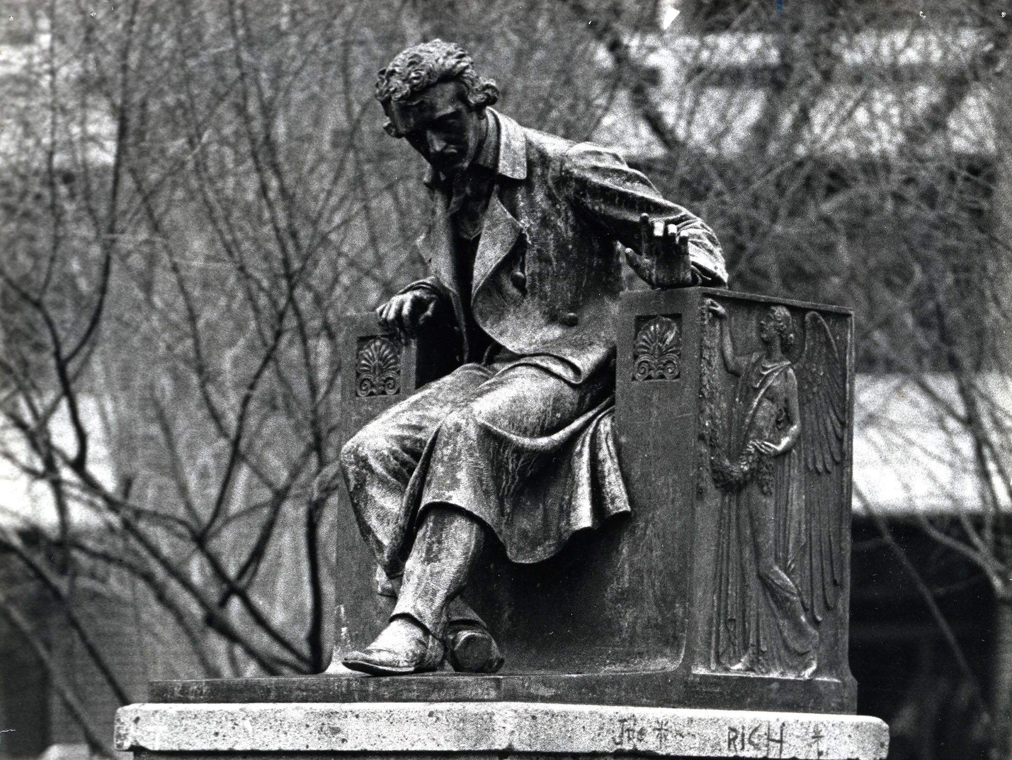Black-and-white photograph of the Edgar Allan Poe statue at the University of Baltimore, depicting Poe seated on a sculpted chair with one arm extended, atop a stone pedestal engraved with his name and dates.