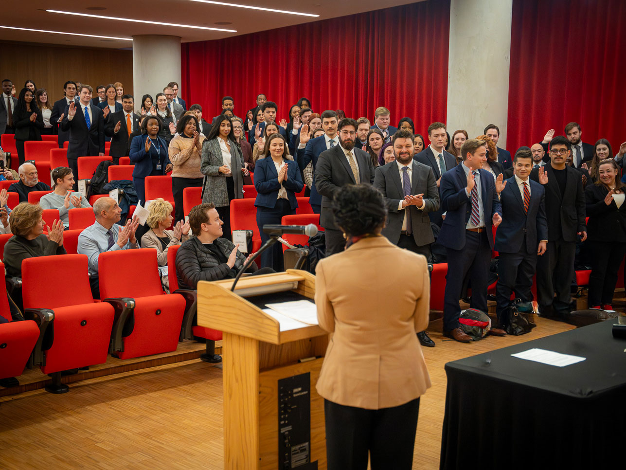 Accessibility description (alt text):  A group of law students stands in a lecture hall with red theater seating, raising their right hands to take an oath. They are dressed in professional attire and face a podium at the front of the room, where a speaker stands reading from prepared remarks. The students fill several rows, with additional attendees seated behind them. The setting appears formal and ceremonial, consistent with a law school swearing in event.