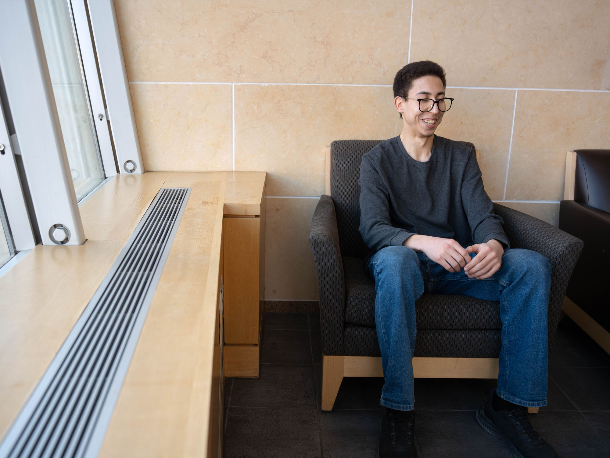 Adam Abidi, a student, sits in a lobby and laughs to himself; the architecture of the building framing his portrait