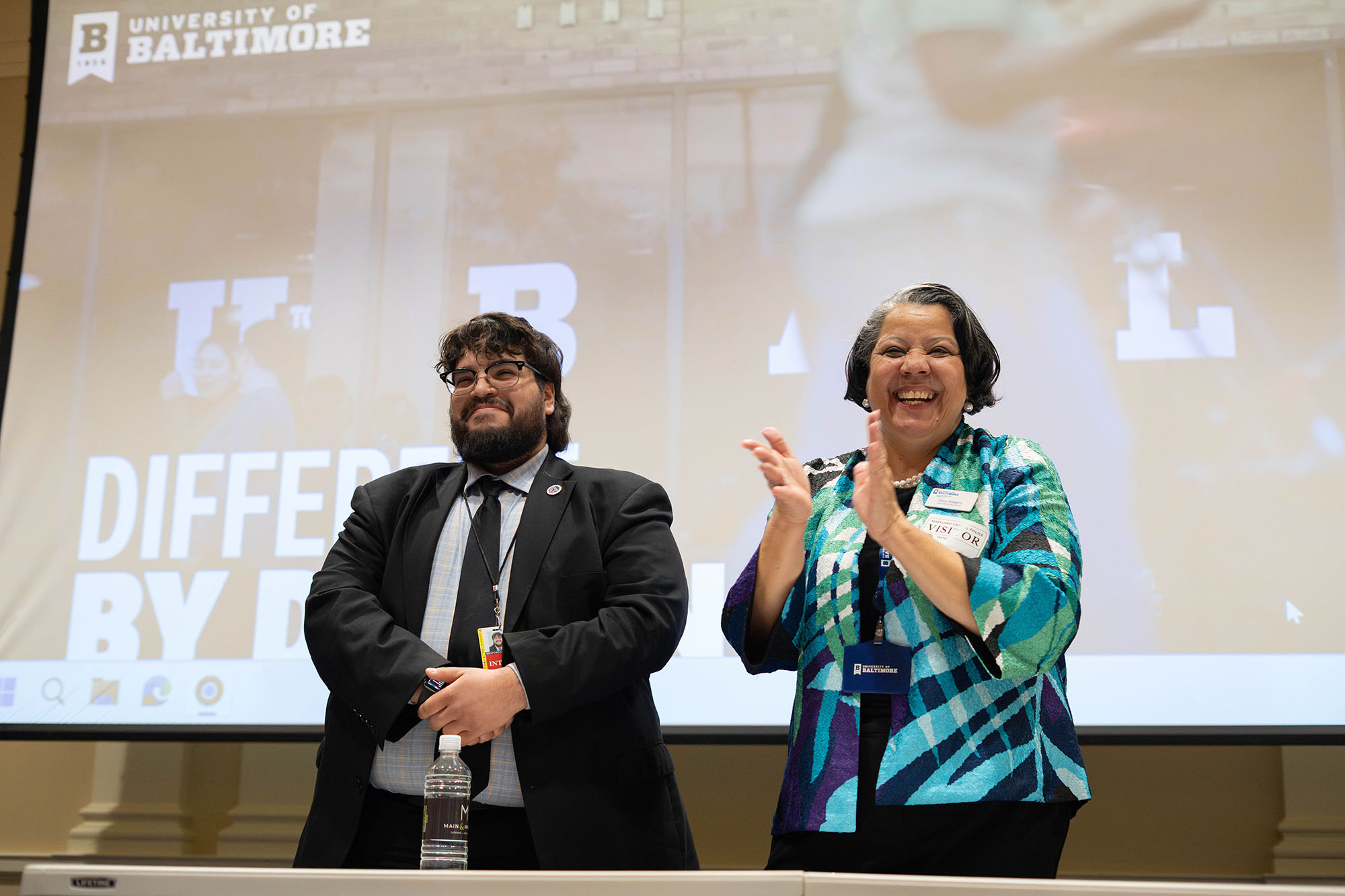 Stacy Rodgers applauds her former student while he looks on. Both stand in front of a presentation screen with the UBalt website featured. 