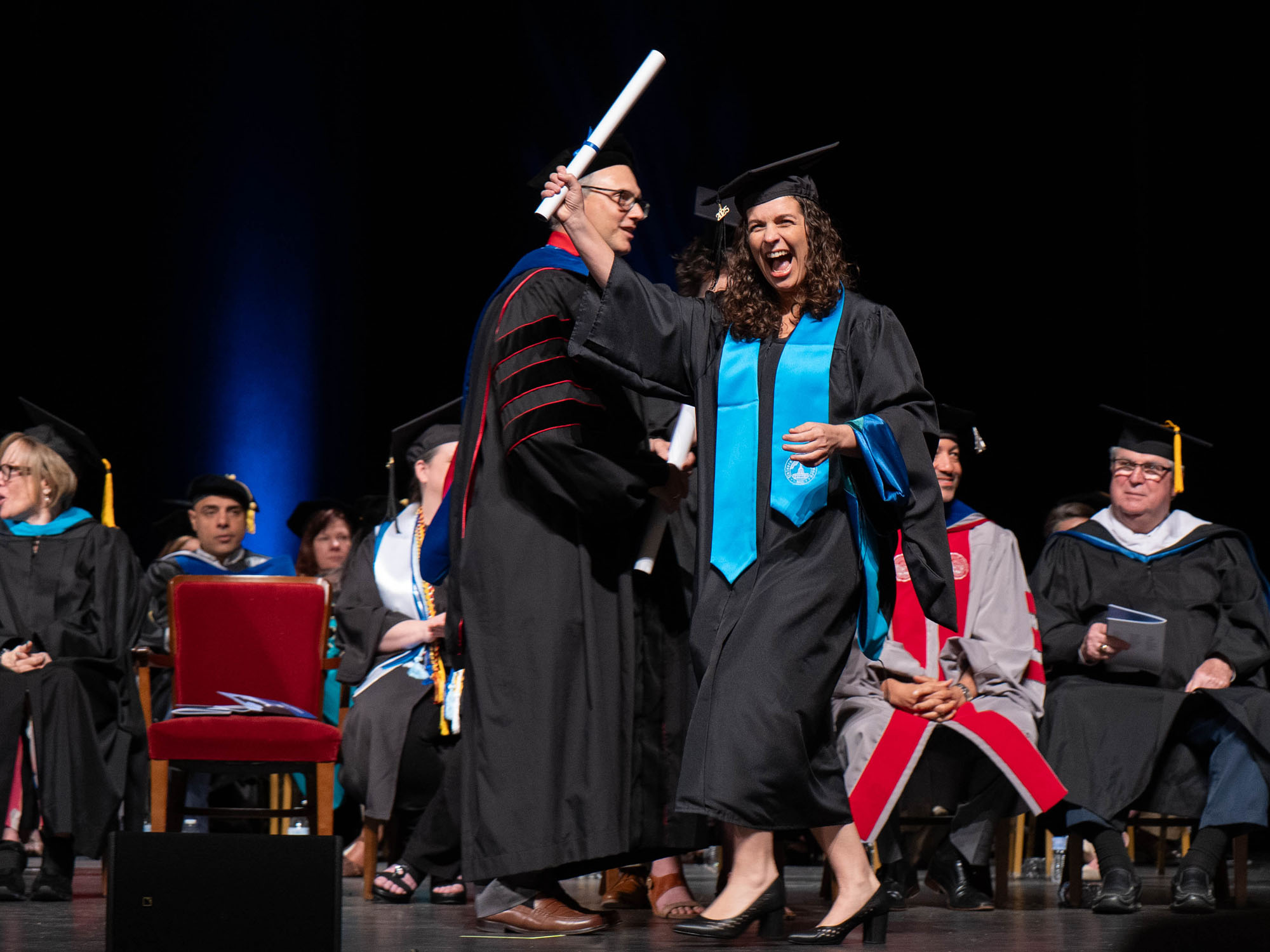 A student waves their diploma while crossing the stage at commencement. 