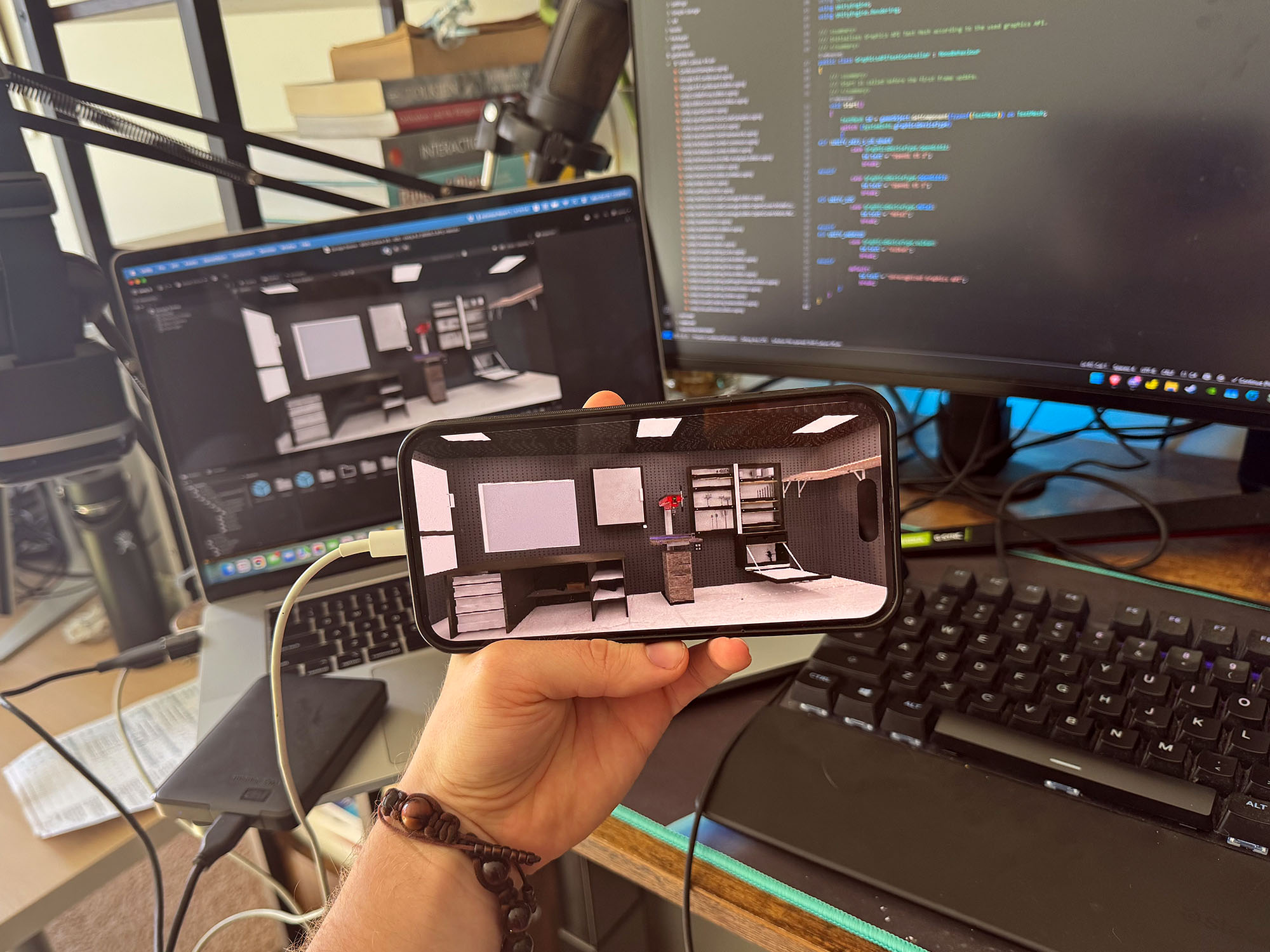 A hand holds up a phone with an image from a video game. In the background are computers showing the game on one screen and the code used to make the game on the other. 