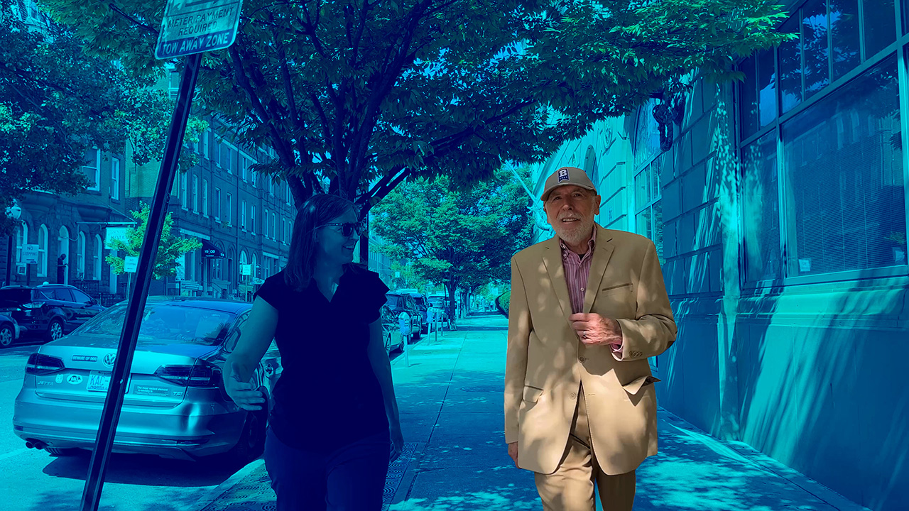 Two people walk on a city sidewalk, coming toward the camera. One, the subject Joe Adler, stands out while the rest of the photo is in a blue hue.
