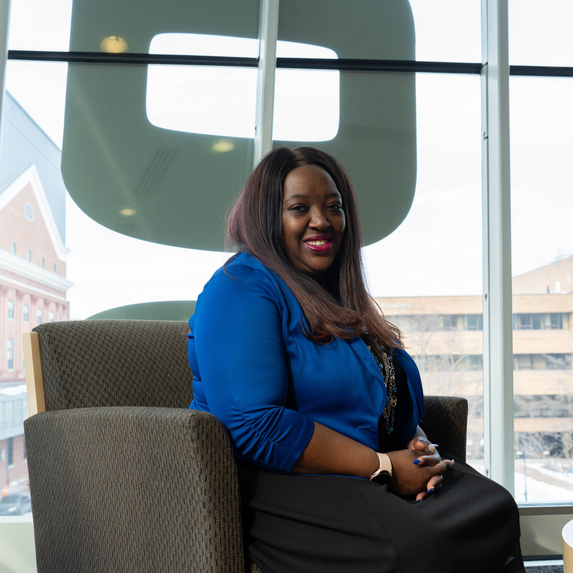 La-Toya Gomez smiles while looking at the camera and sitting in the Student Center, in front of a big window.