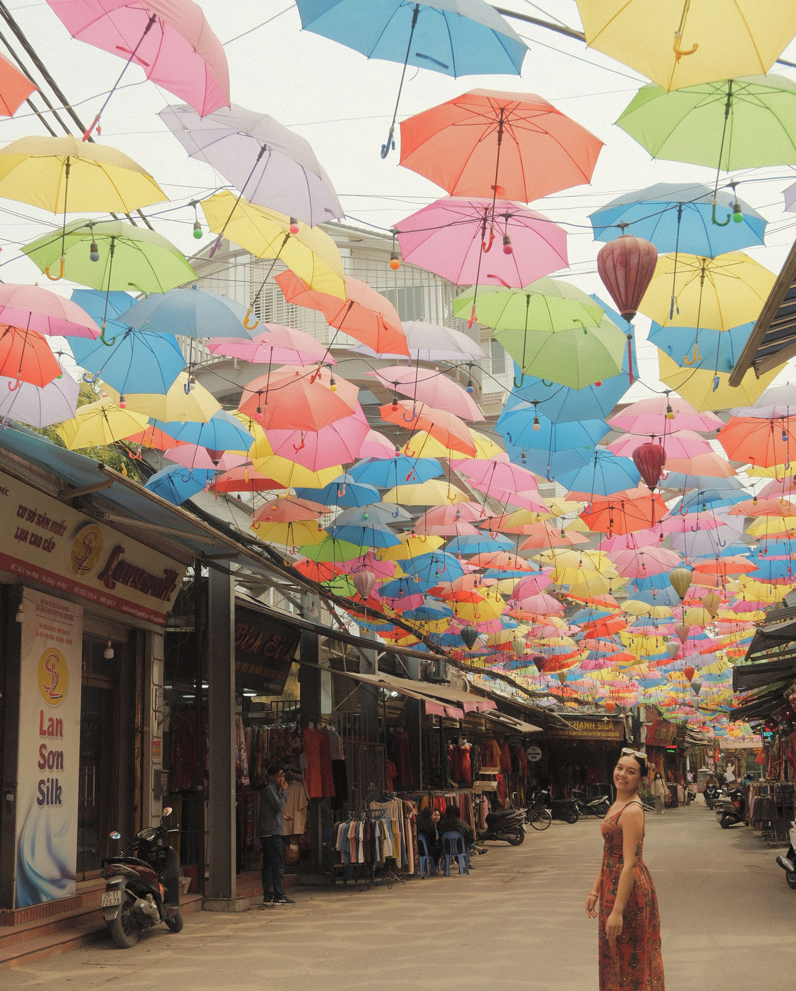 A woman stands in an open market in Vietnam below a ceiling of colorful umbrellas all hanging in rows.