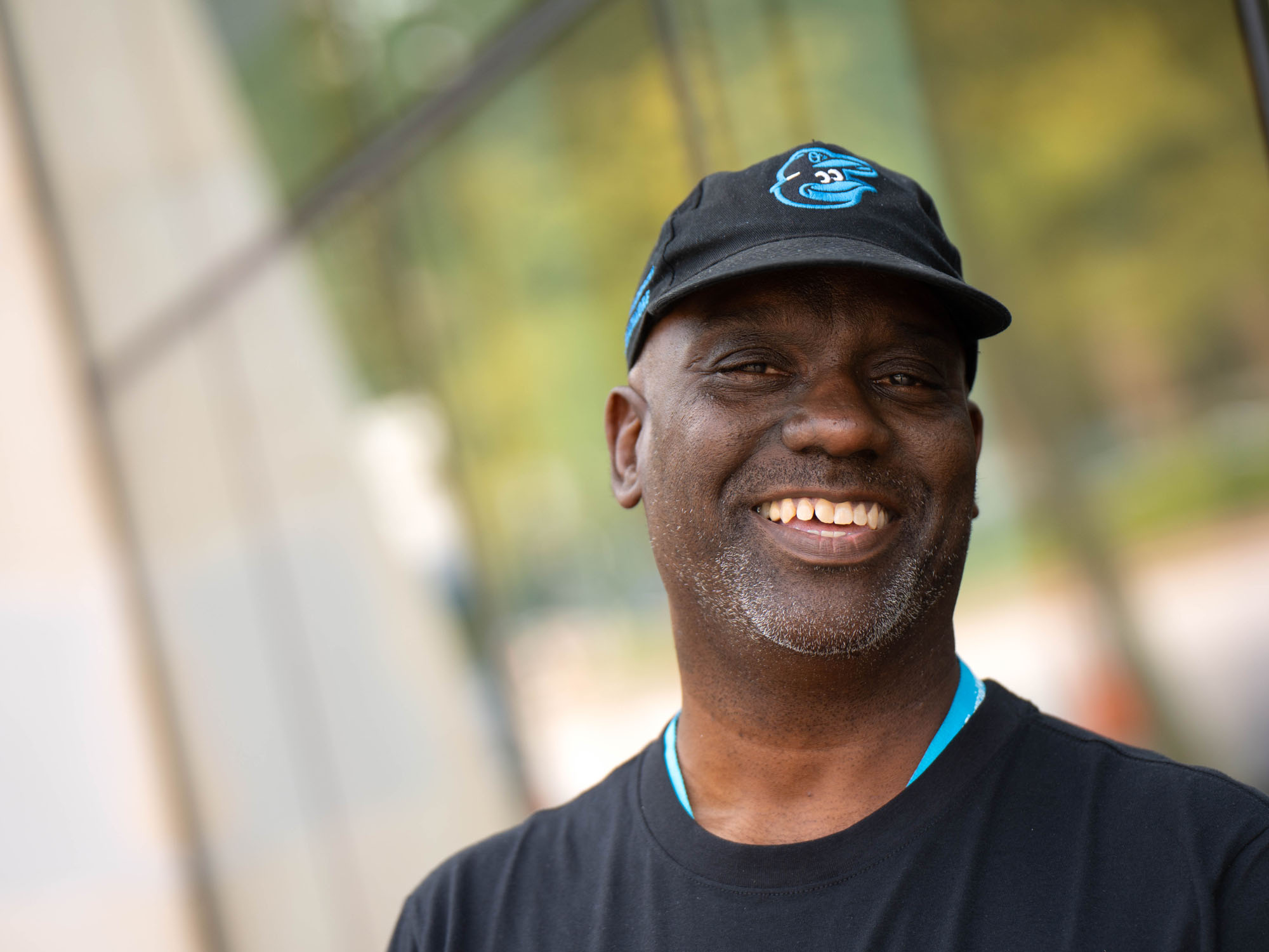 Diarra, a student at The University of Baltimore, smiles while wearing an Orioles ballcap and standing outside a campus building.