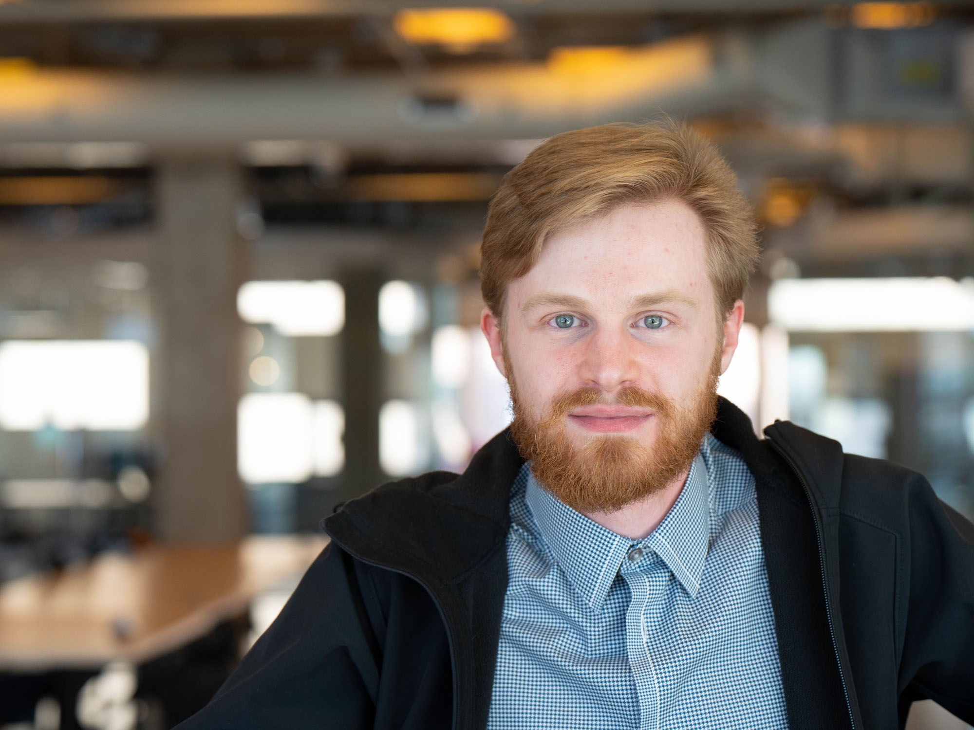 A student wearing a jacket and button-down shirt smiles in a portrait shot in the University of Baltimore library.