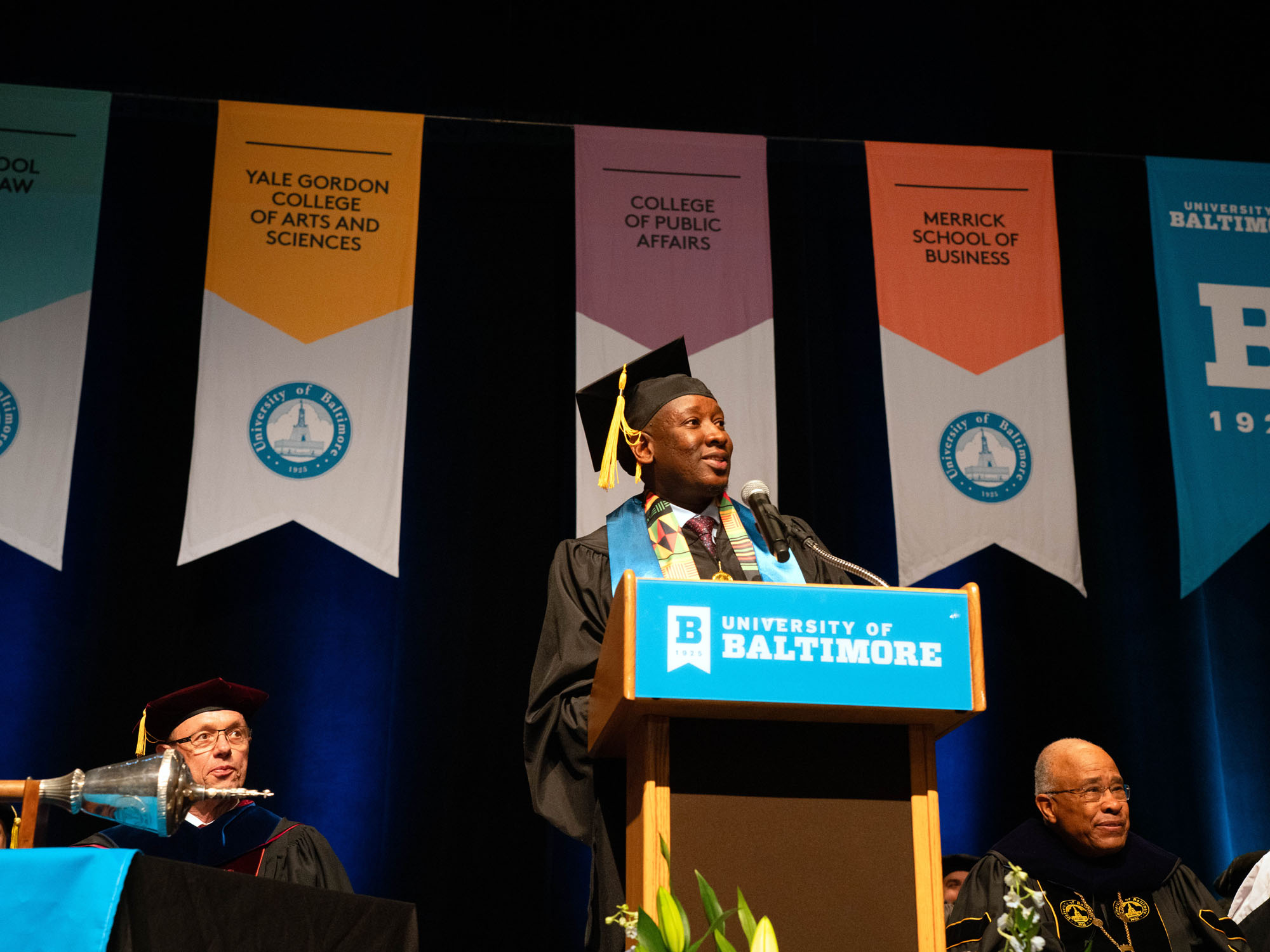 Erick Masaua, in his cap and gown, speaks from the podium on stage during commencement.