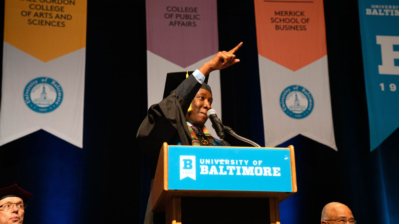 Erick Masaua points toward his family while giving his commencement speech on stage in his cap and gown.