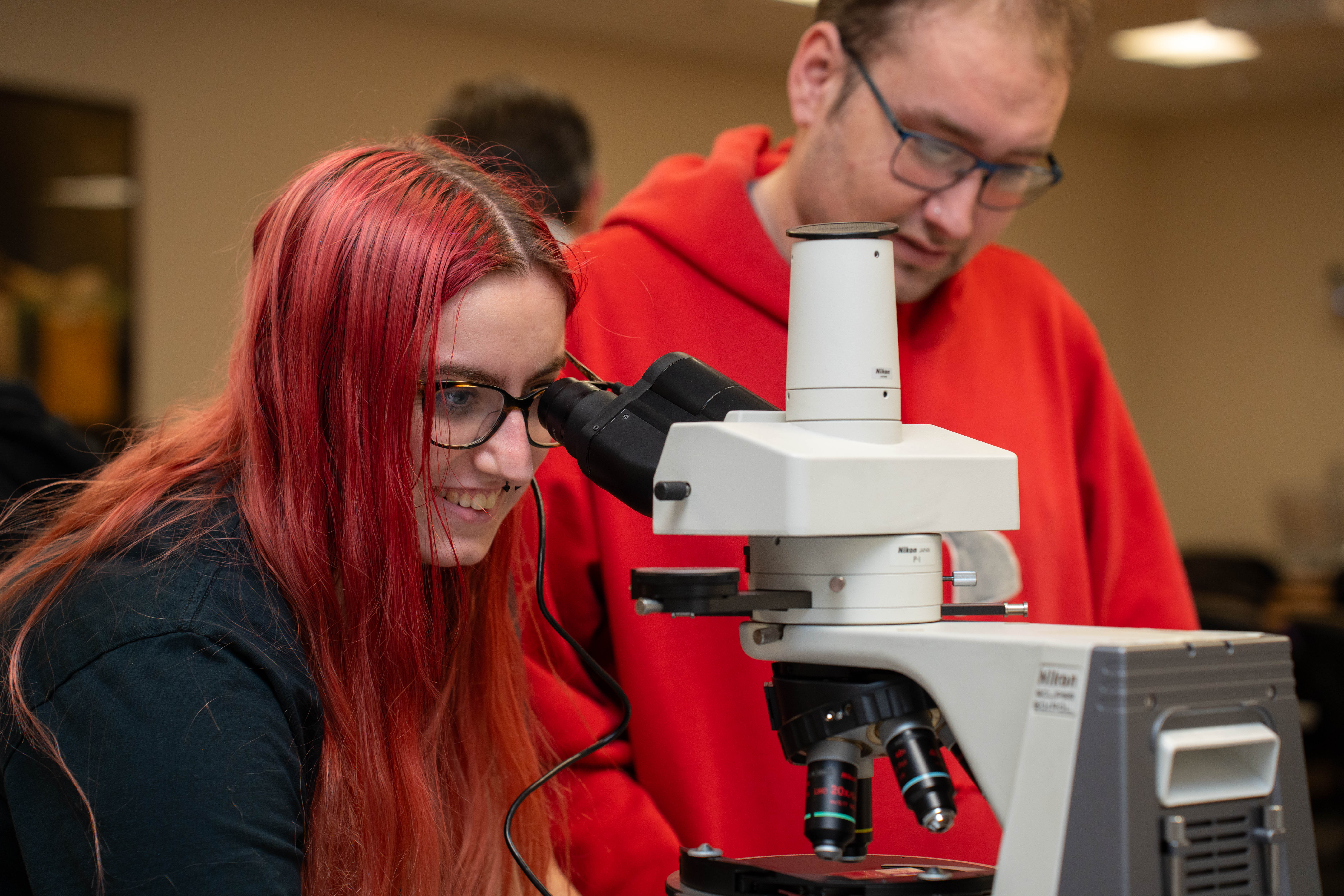 A student smiles while looking inside a microscope while their lab partner looks on. 