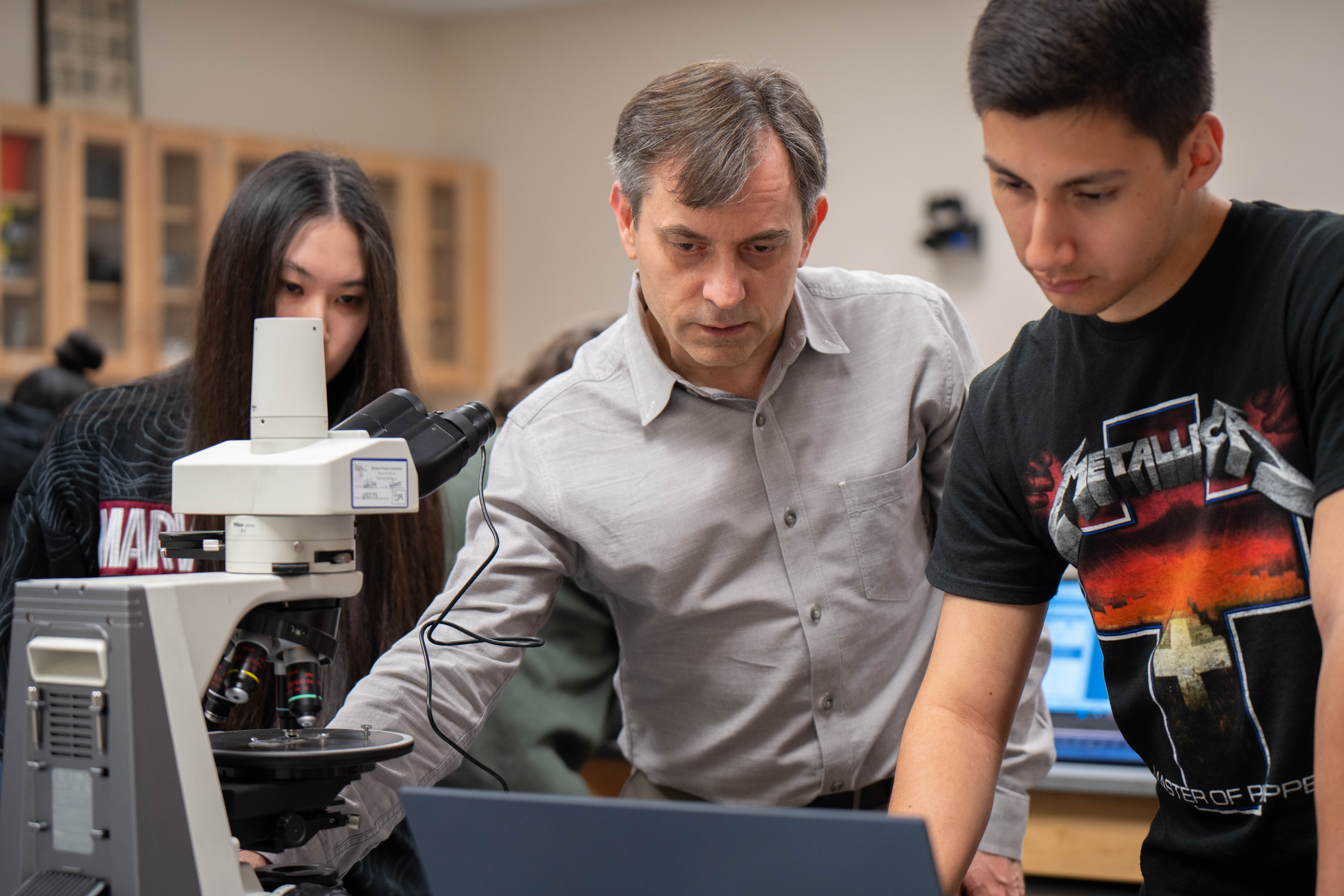 A professor looks over a laptop screen with a student as that professor adjusts the microscope.
