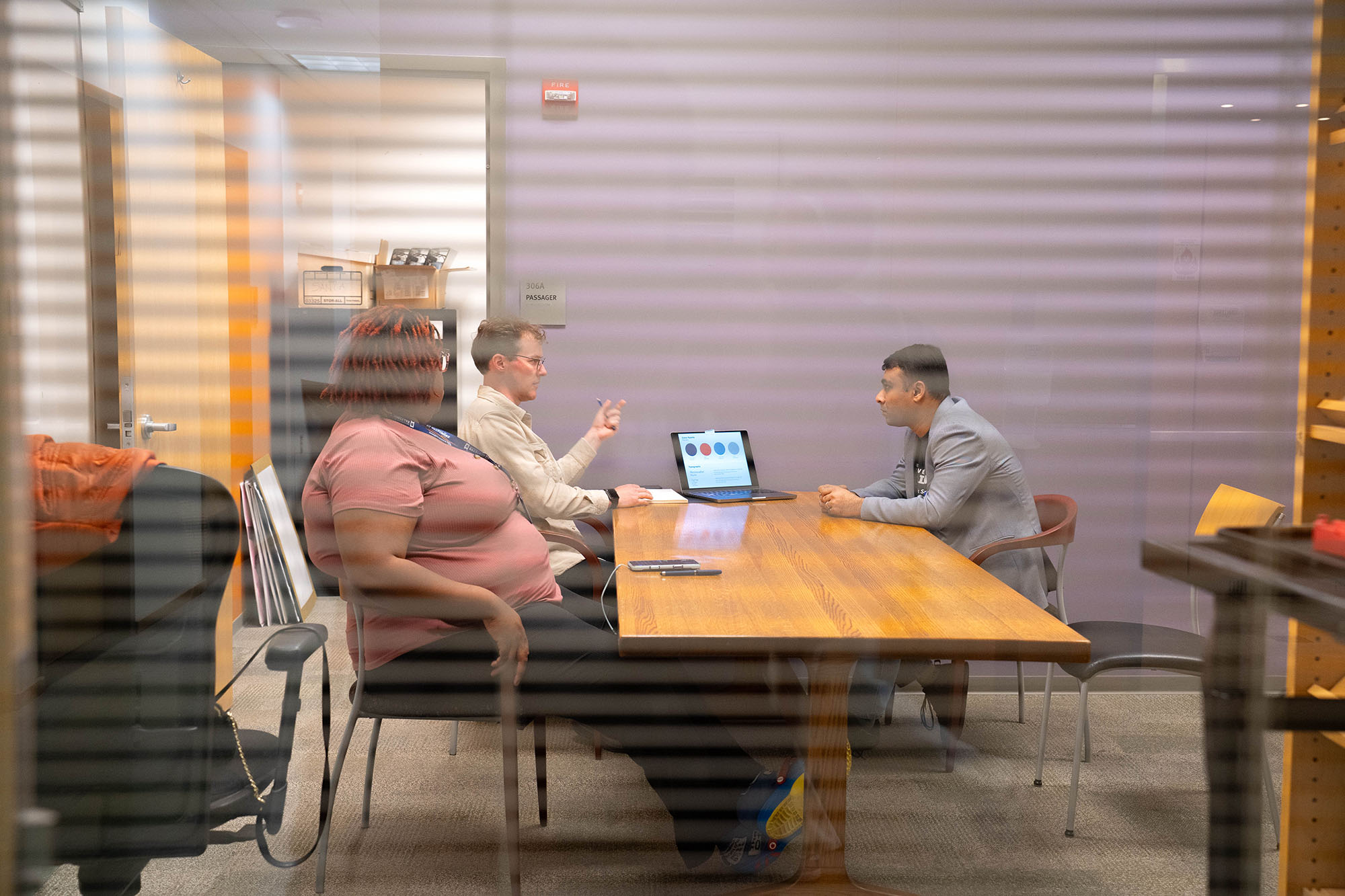 A student group working at a table is captured through the blinds of a window. 
