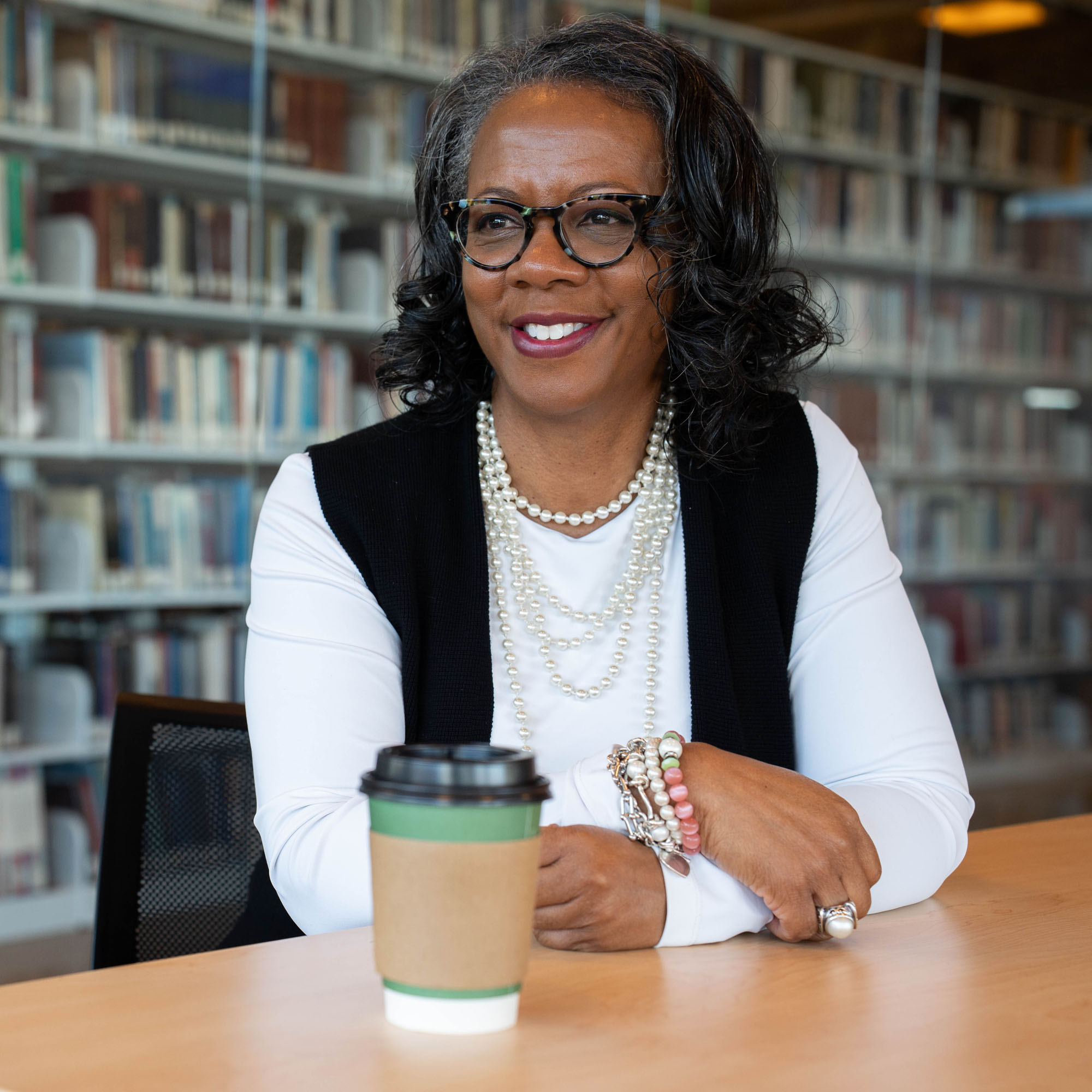 Jan Williams sits at a table with a coffee cup looking on. 
