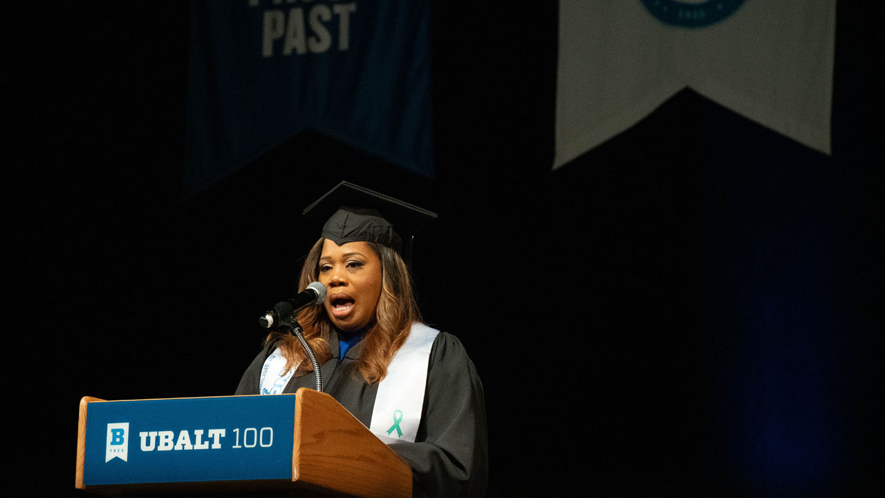 A student in cap and gown address graduates from a podium. 