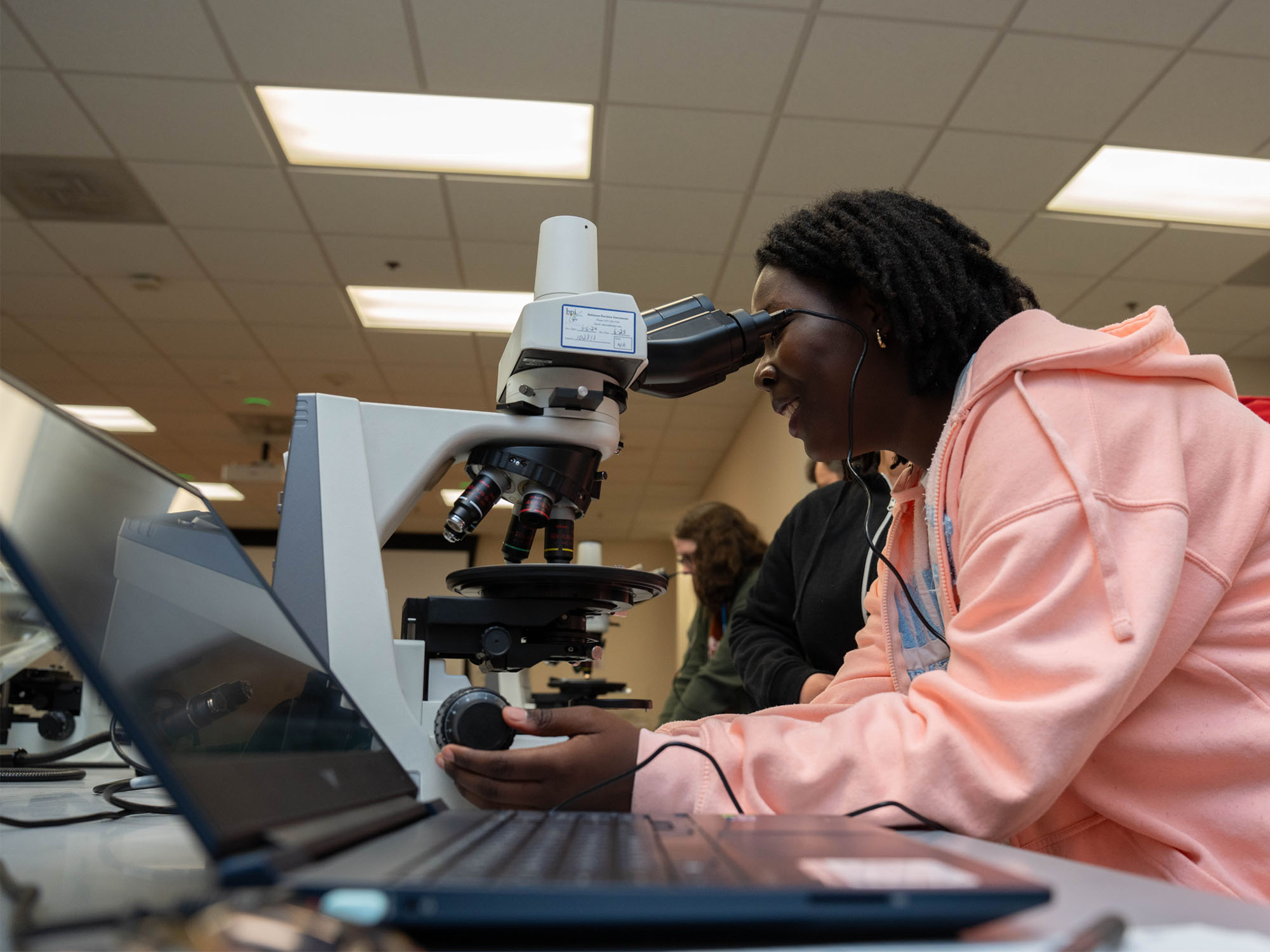 A student looks into a microscope with a laptop in the foreground.