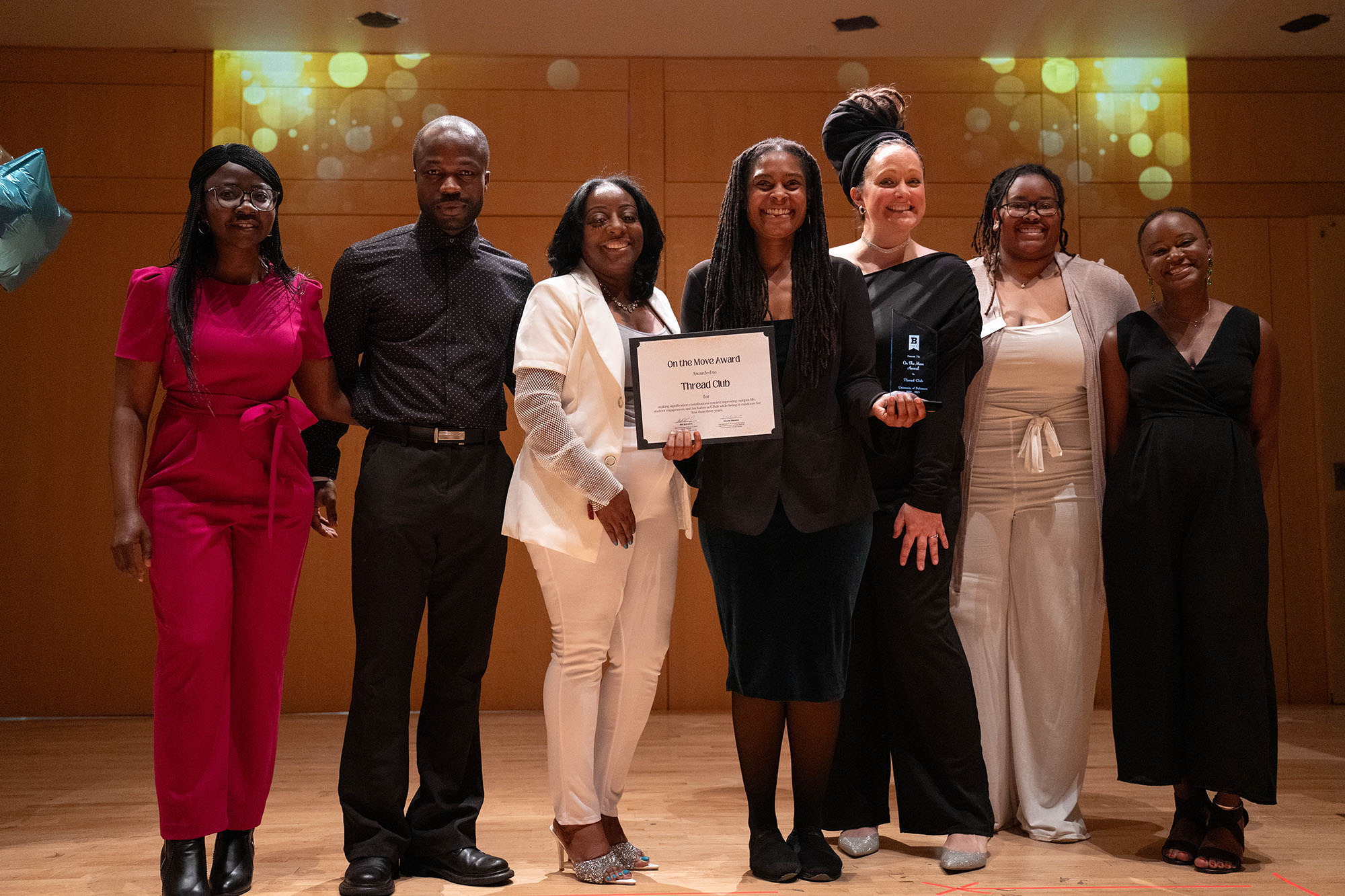 Seven students stand on a stage, the one at center holding two awards.