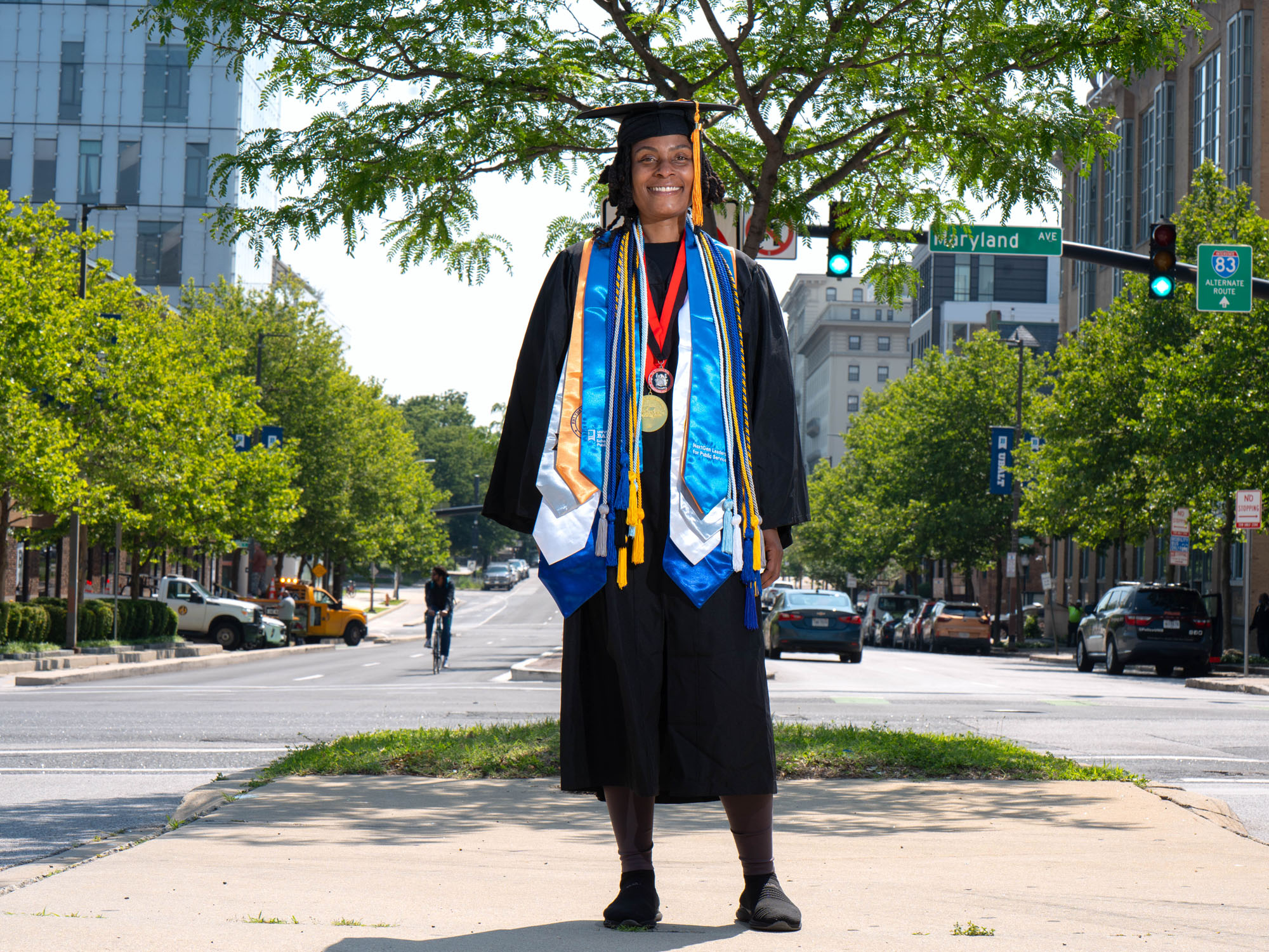 A graduate in cap, gown, stoles and cords stands on a road median with the campus in the background.