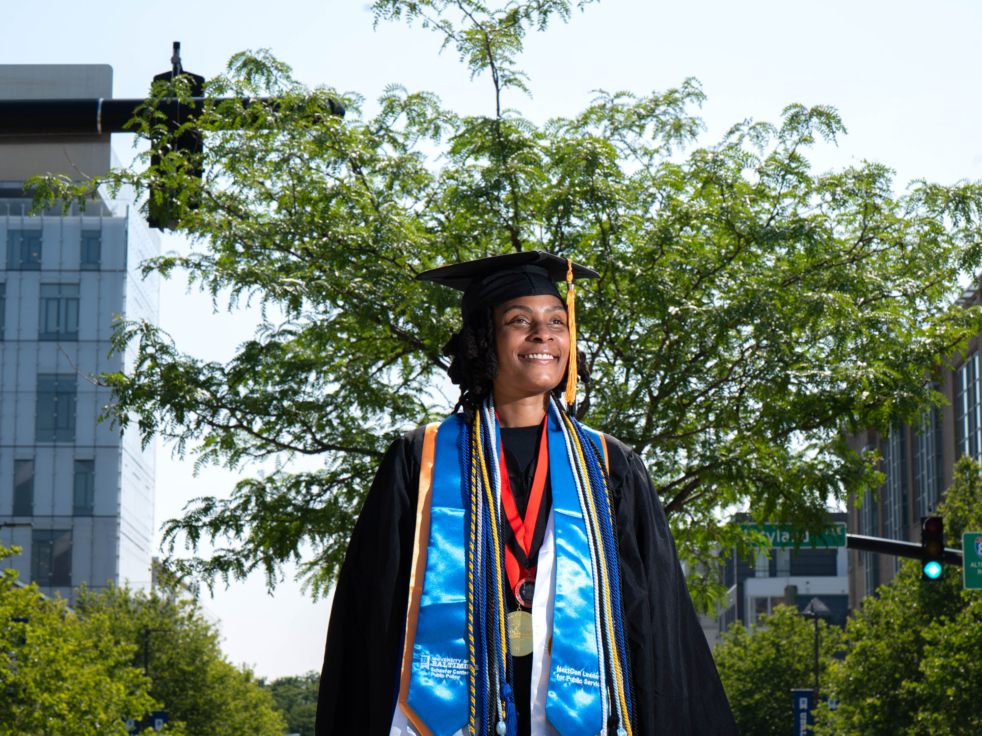 A graduate in cap, gown, stoles and cords stands on a road median with the campus in the background.