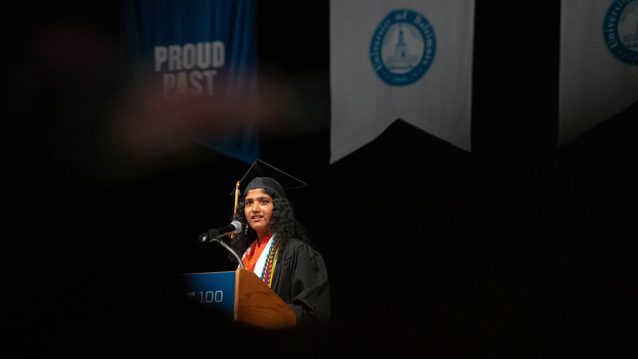A speaker is seen on stage through gaps around grad caps.