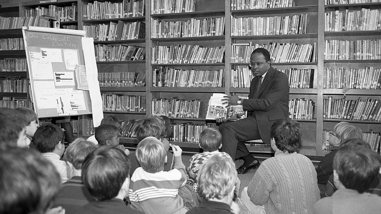 In an older black and white image, Kurt Schmoke, then mayor of Baltimore, reads to children in a library.