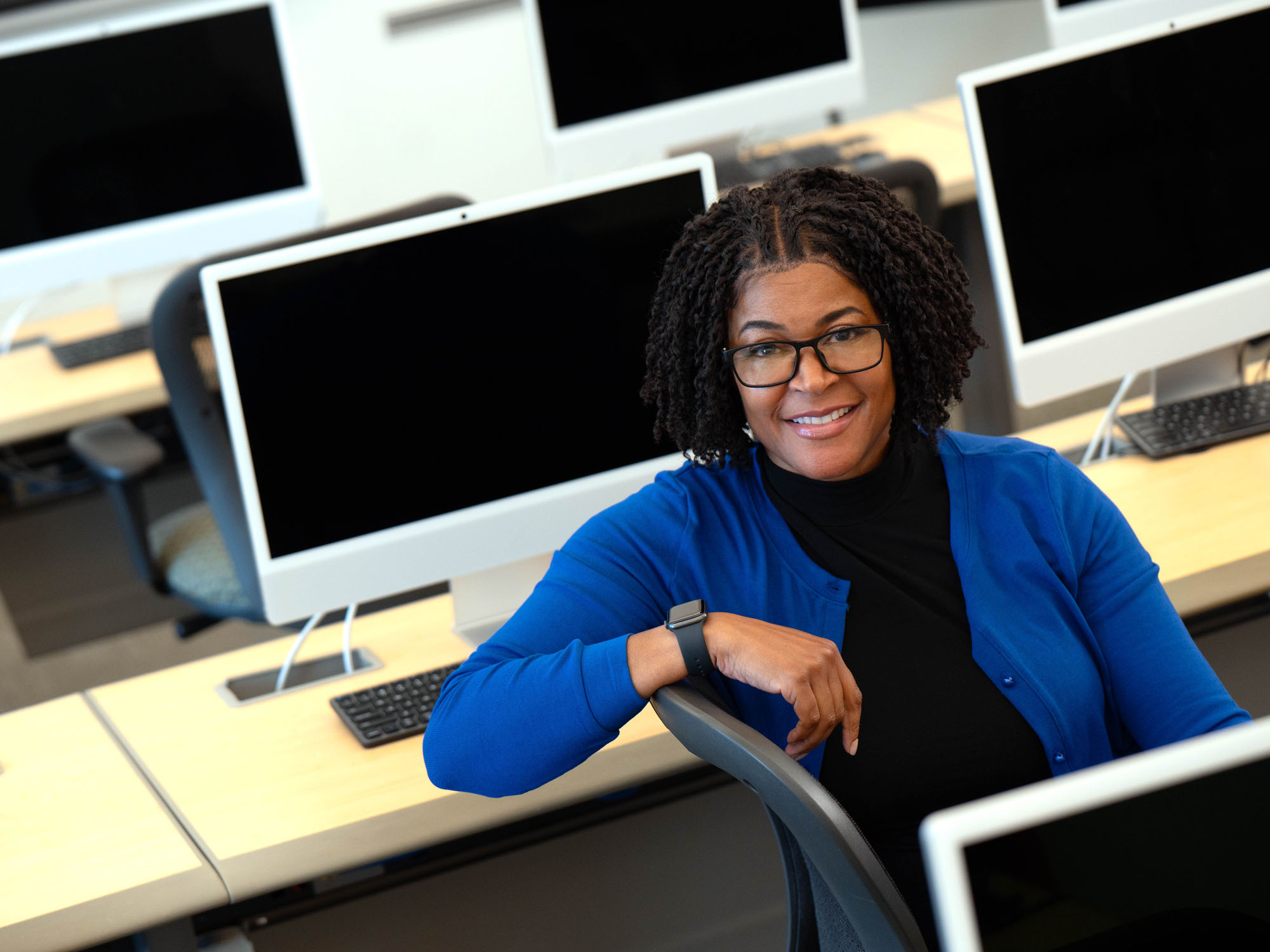A student sits casually in a computer lab and smiles at the camera during a portrait shoot. 