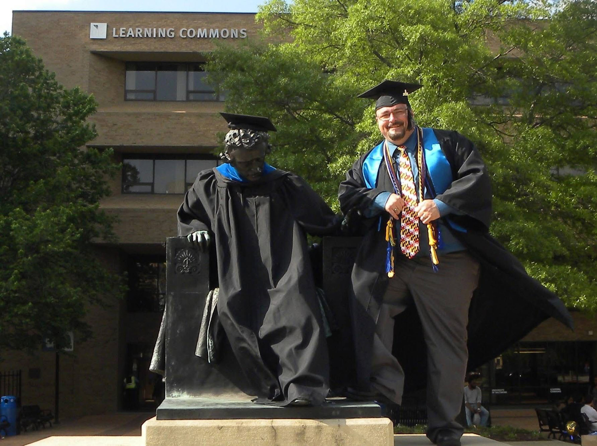 In his cap and gown, newly minted alum Troy Pritt poses with the Edgar Allan Poe statue on UBalt's campus.