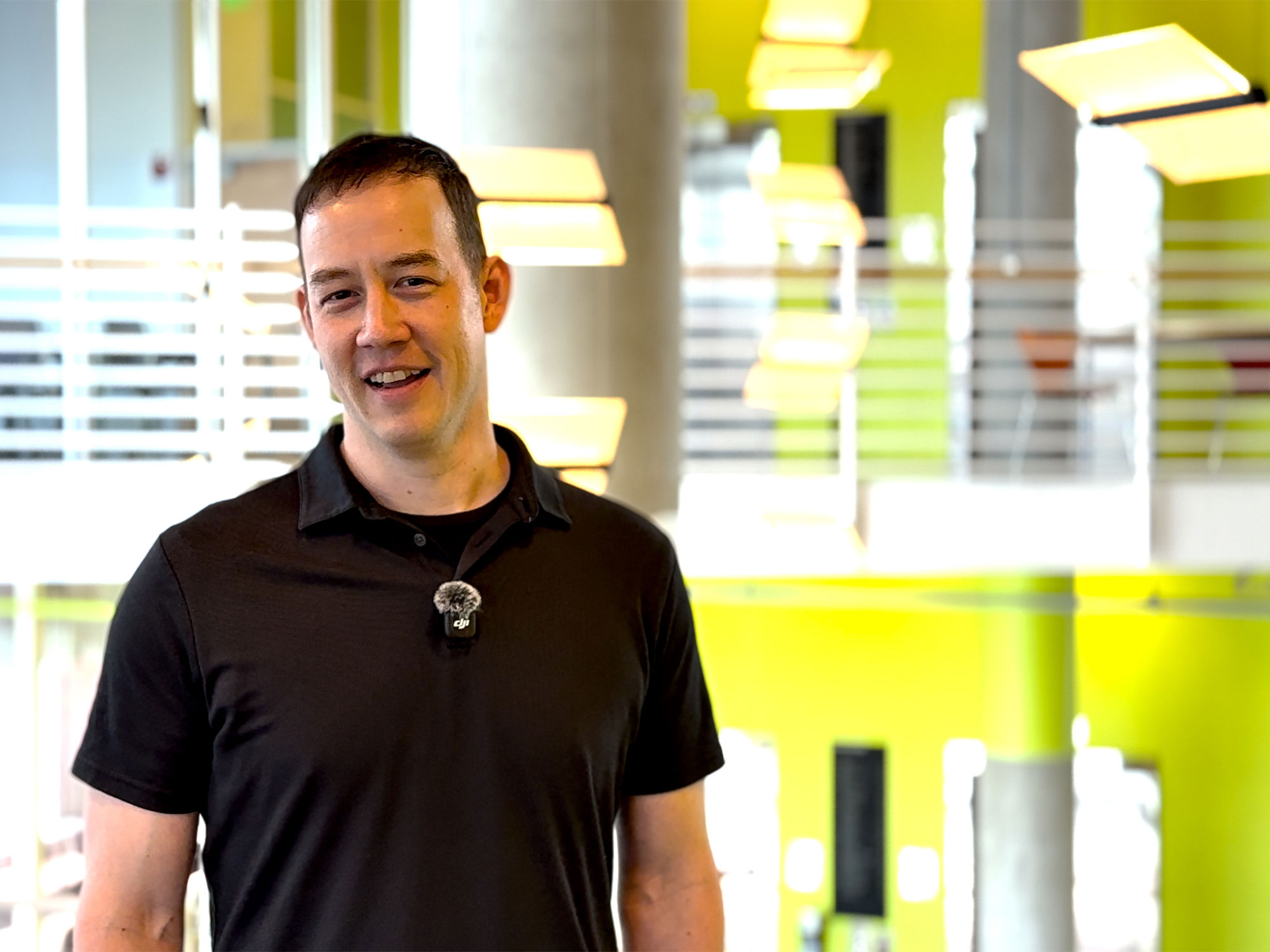 James Pollock smiles while standing in a bright, modern interior with glass walls, lime-green accents and suspended lights. He wears a black polo shirt with a wireless microphone clipped to his chest.