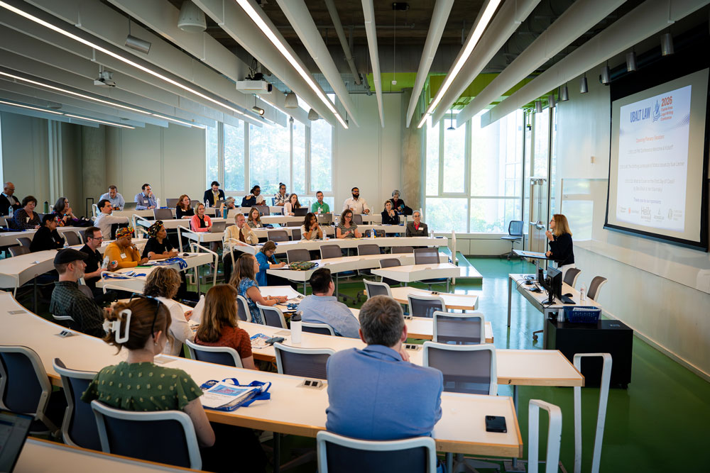 Rows of seated conference attendees in a tiered lecture hall, facing a presenter and projection screen.