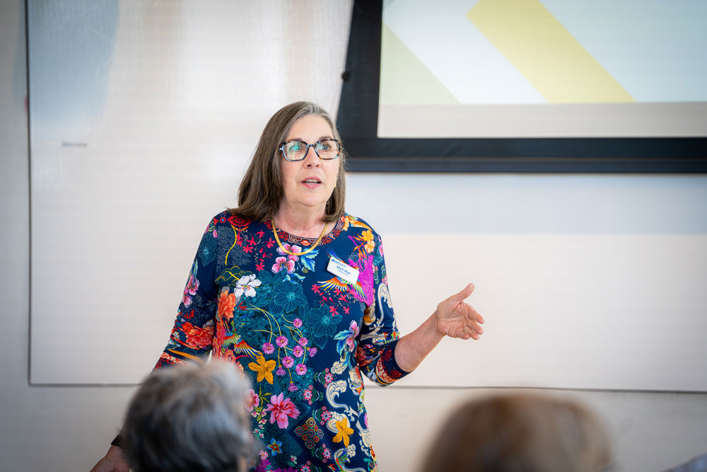 A woman speaking to an audience, standing in front of a presentation screen with one arm raised."