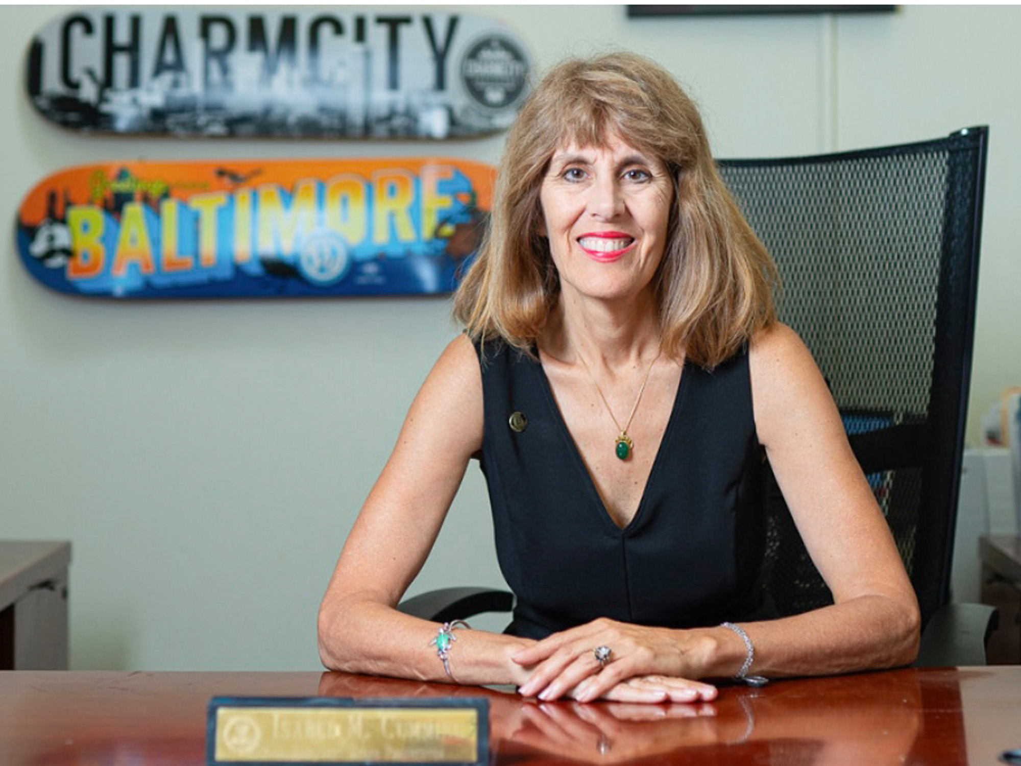 Photo of a smiling woman with shoulder-length light brown hair standing at a wooden desk in an office, wearing a black sleeveless top with a V-neck and a gold necklace with a green pendant. She rests her hands on the desk in front of a name plate. On the wall behind her are two decorative skateboard decks, one reading 