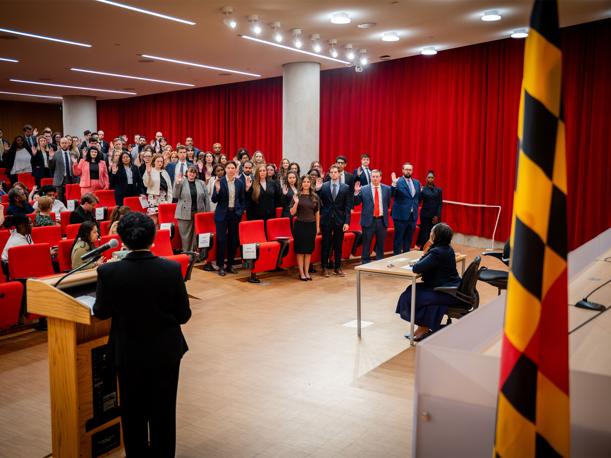 UBalt Law students take an oath during a ceremony in an auditorium, with the Maryland state flag in the foreground.