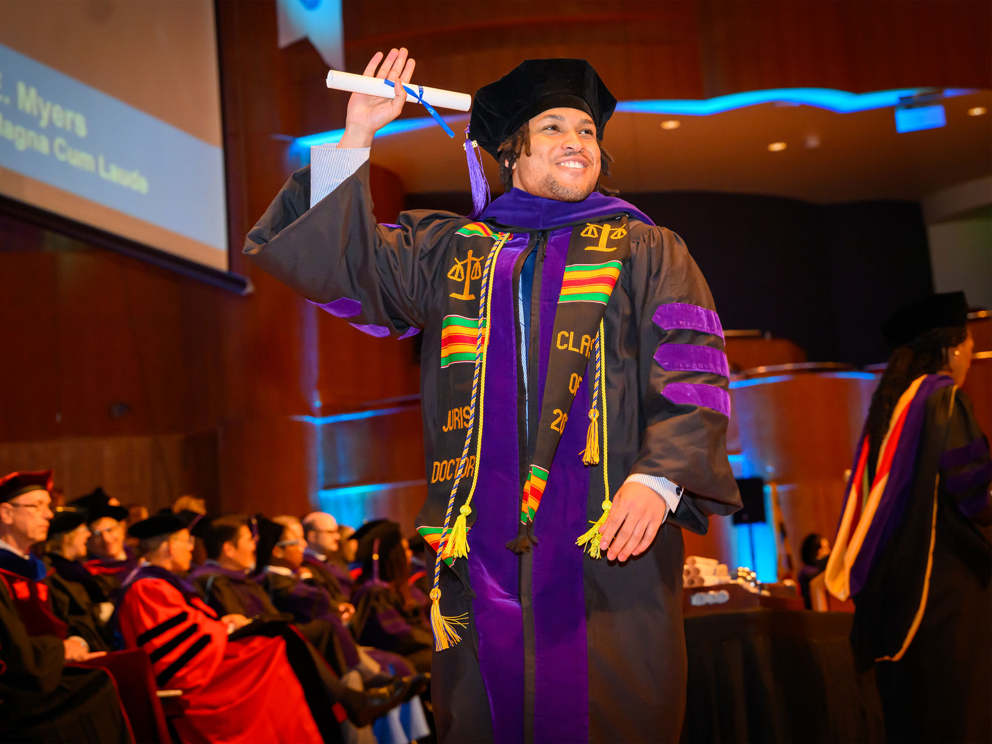 A UBalt Law graduate smiles and raises a diploma while walking across the stage during commencement.
