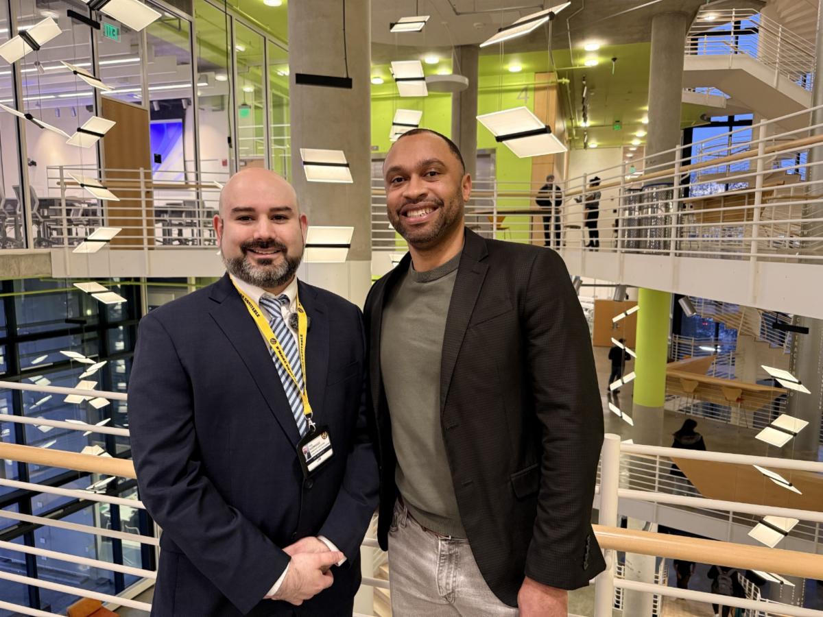 Paul Rodriguez and Stephen Cirino stand together in a bright, modern atrium at the University of Baltimore School of Law. Both are smiling and dressed in professional attire, reflecting a welcoming and collegial environment. The contemporary space, with open balconies and suspended light installations, highlights the school’s vibrant academic community and spirit of connection.