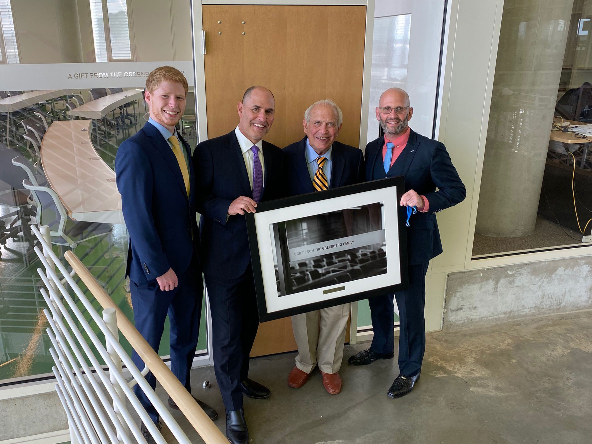 Four members of the Greenberg family stand together inside UBalt Law, holding a framed photo commemorating a family gift.
