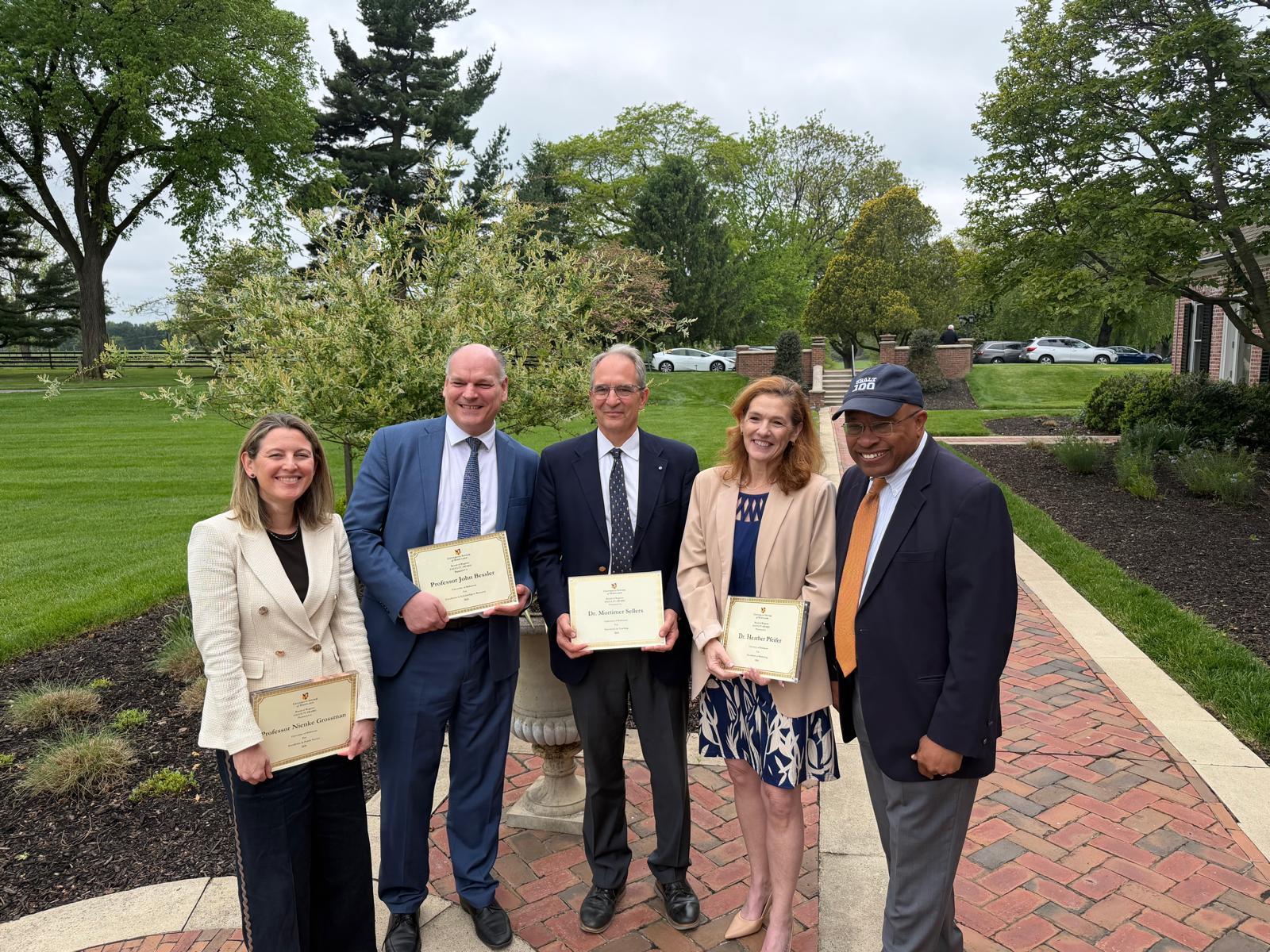 Five individuals stand outdoors on a brick pathway holding award certificates and smiling for a group photo. The group is surrounded by landscaped gardens, green lawns, and mature trees on a cloudy day.