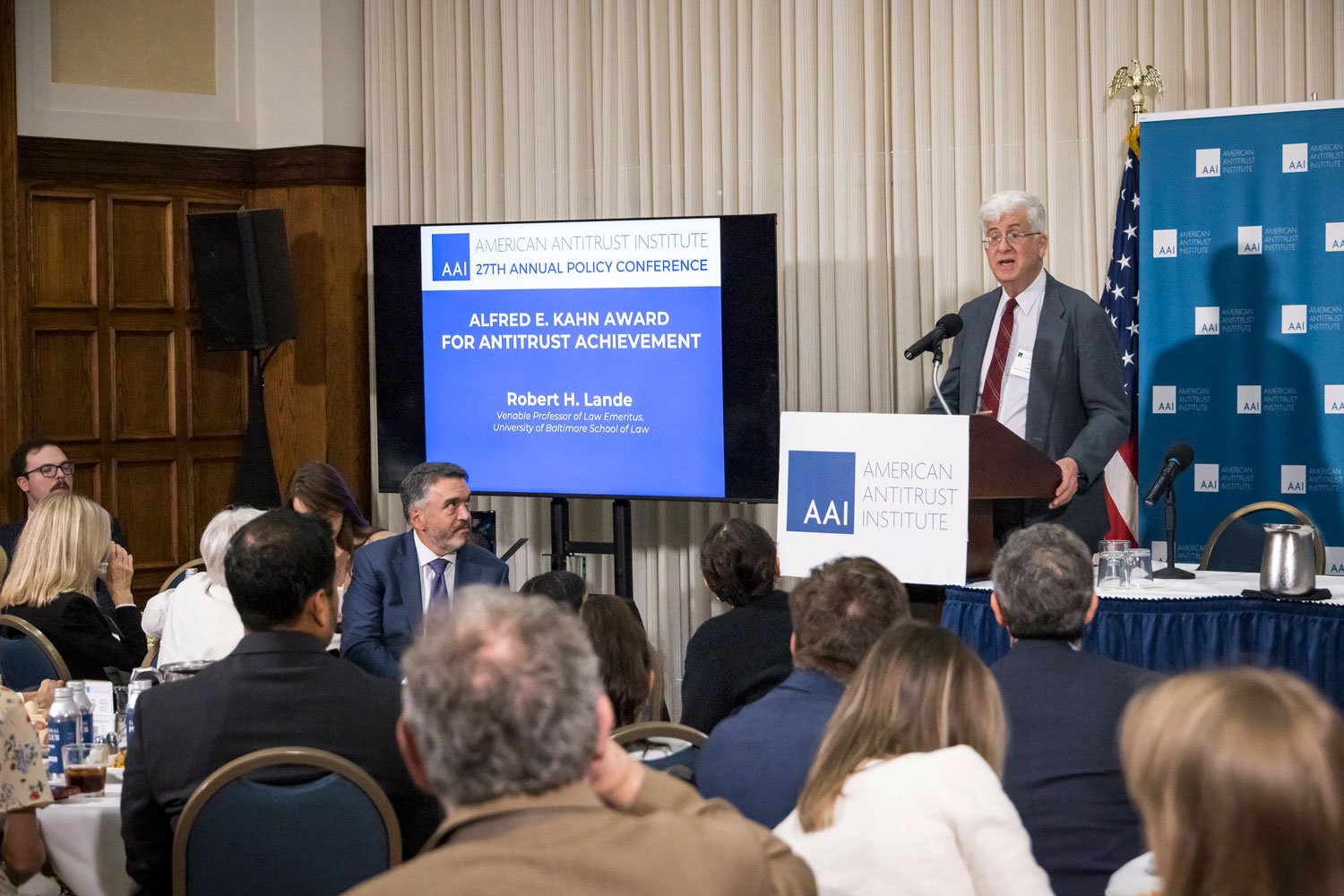 Robert H. Lande speaks at a podium while receiving the Alfred E. Kahn Award for Antitrust Achievement during the American Antitrust Institute’s 27th Annual Policy Conference. Attendees sit at tables facing the stage.