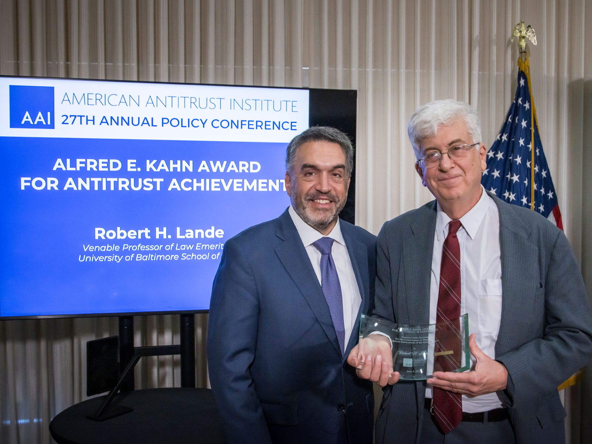 Robert H. Lande accepts the Alfred E. Kahn Award for Antitrust Achievement at the American Antitrust Institute's 27th Annual Policy Conference, standing with the presenter while holding the award in front of a conference display and a U.S. flag.