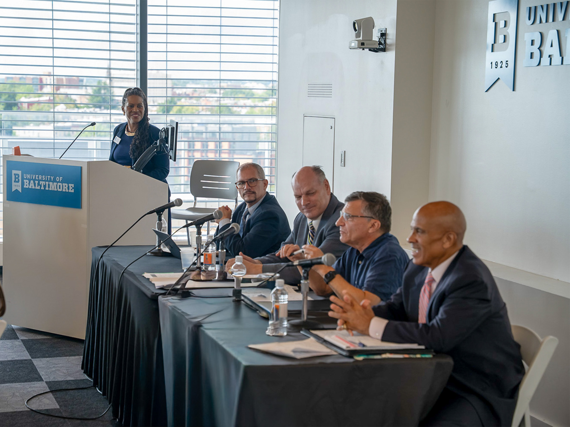 A woman speaks at a University of Baltimore podium beside four panelists seated at a table with microphones during an event. Audience members sit in the foreground.