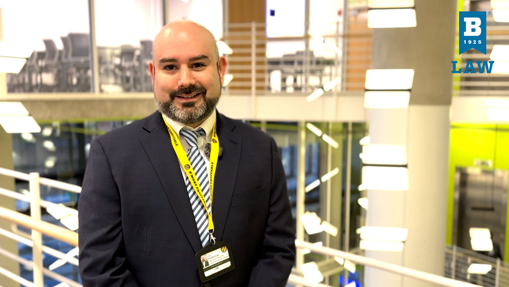 Paul Rodriguez, wearing a dark suit, striped tie, and University of Baltimore School of Law lanyard, stands smiling in a bright, modern atrium. He faces the camera with hands together, appearing confident and professional. The background shows open railings, glass walls, and suspended lights in a contemporary academic building.