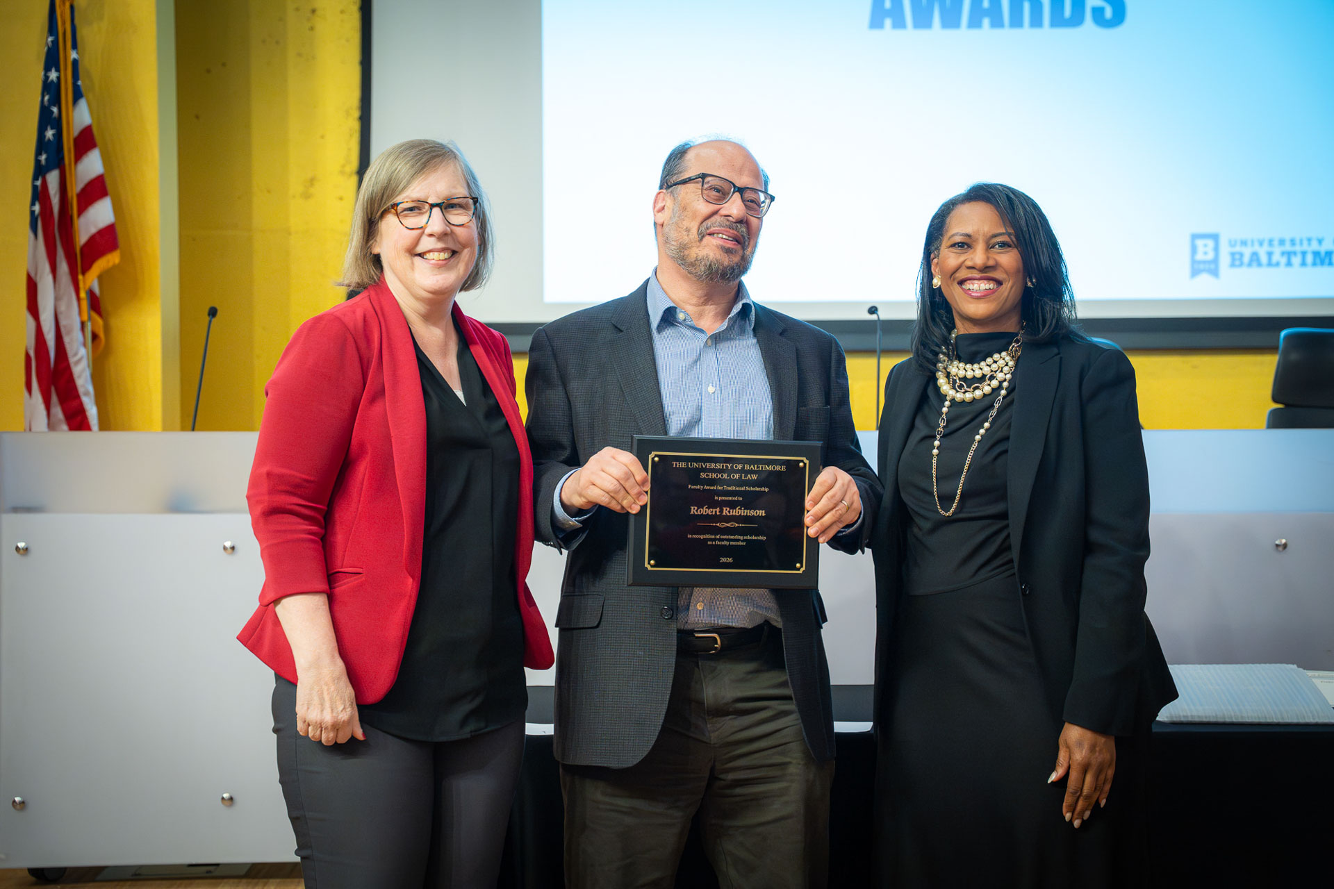 **Alt text:** Three individuals stand together at an awards ceremony, with the person in the center holding a recognition plaque. They pose in front of a presentation screen and an American flag inside a lecture hall or auditorium. The group is smiling during the award presentation.