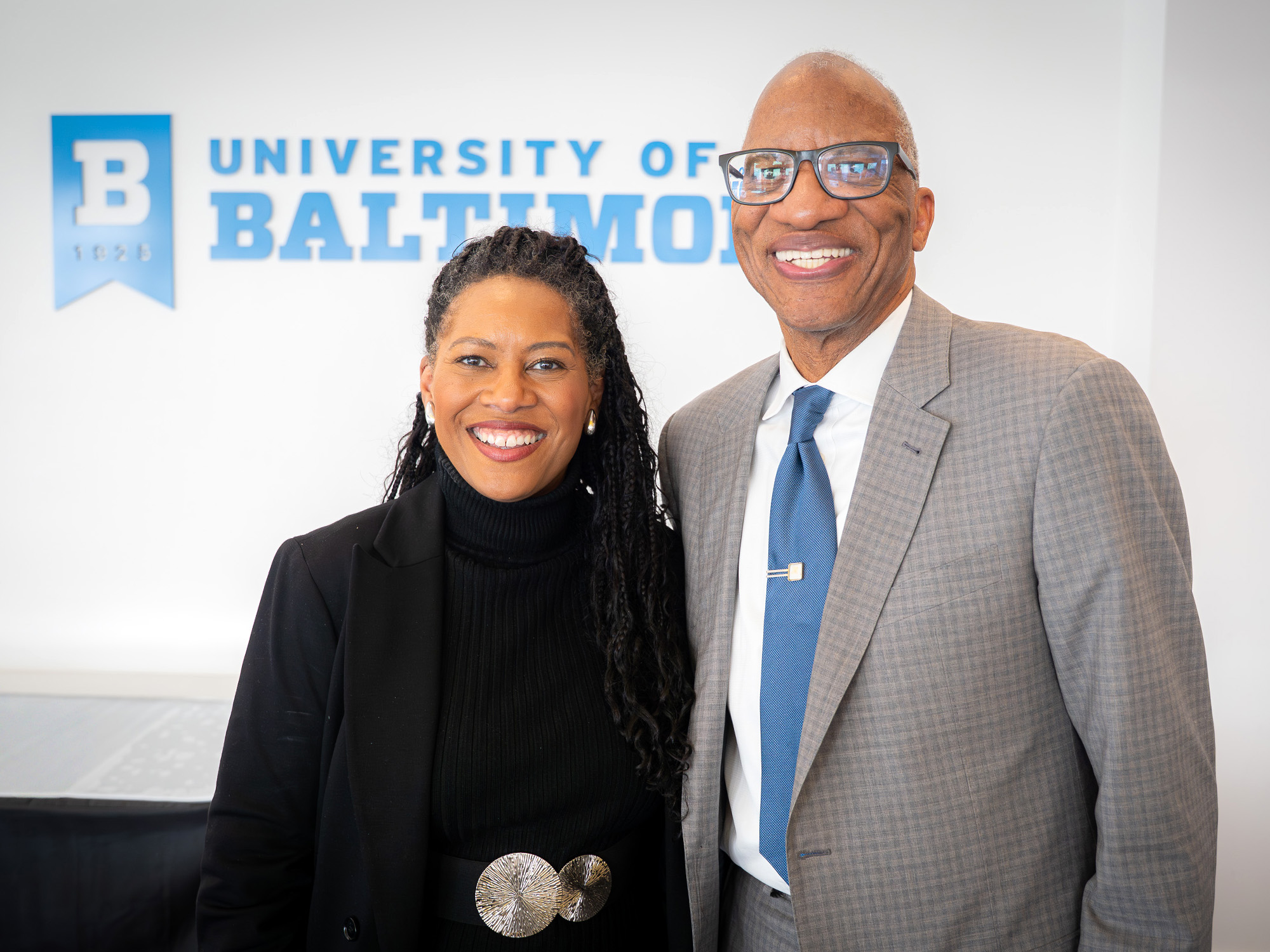 Two people standing together and smiling in front of a University of Baltimore sign. Both are dressed in professional attire, posing for a photo at the University of Baltimore School of Law.