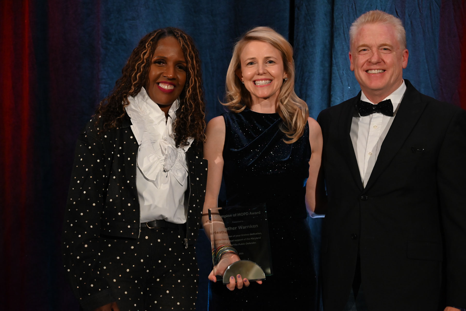 Three people pose together at an awards event, with one recipient holding an award plaque.