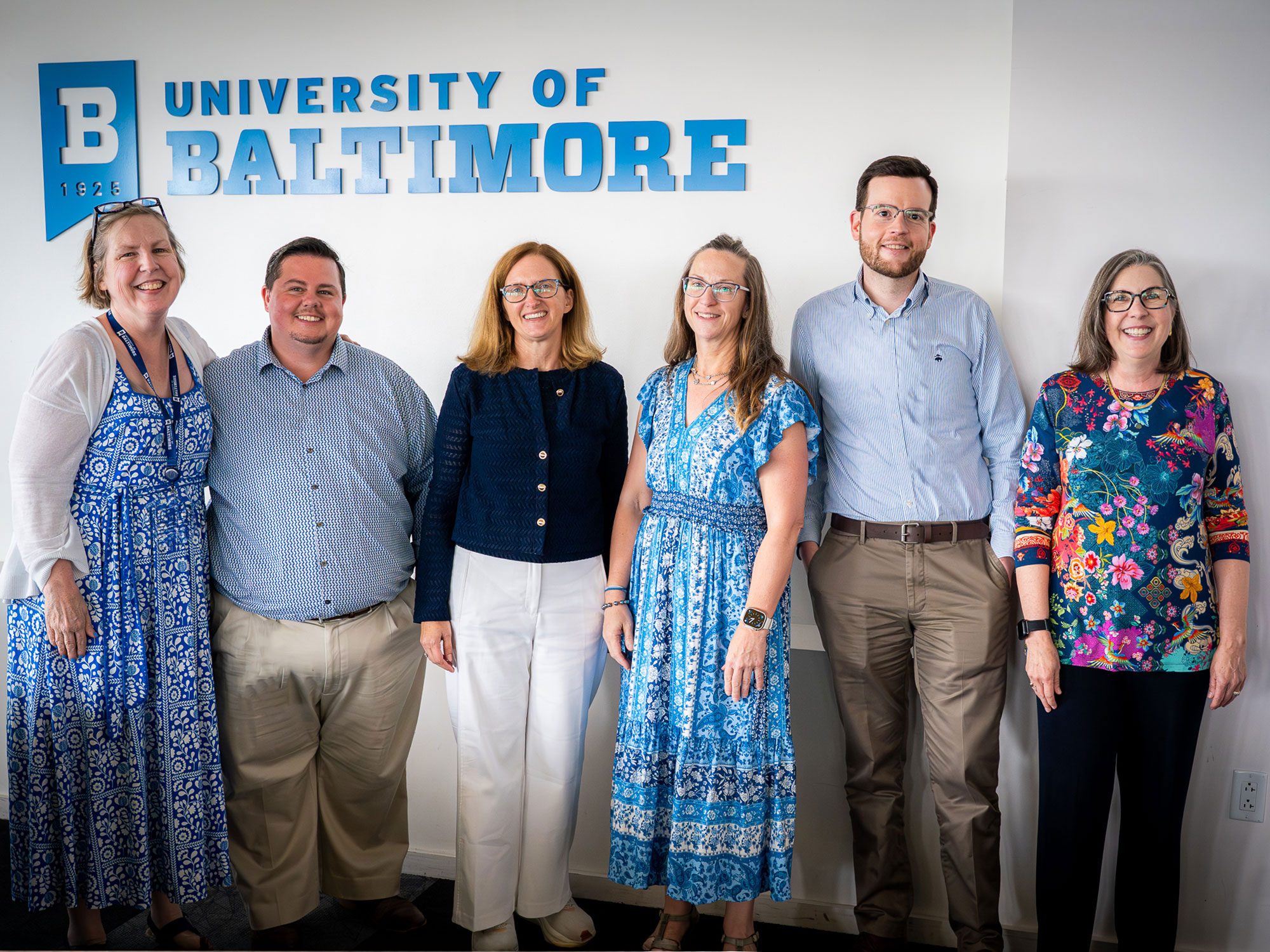 Six people stand smiling in front of a University of Baltimore wall sign, dressed professionally in business casual attire.