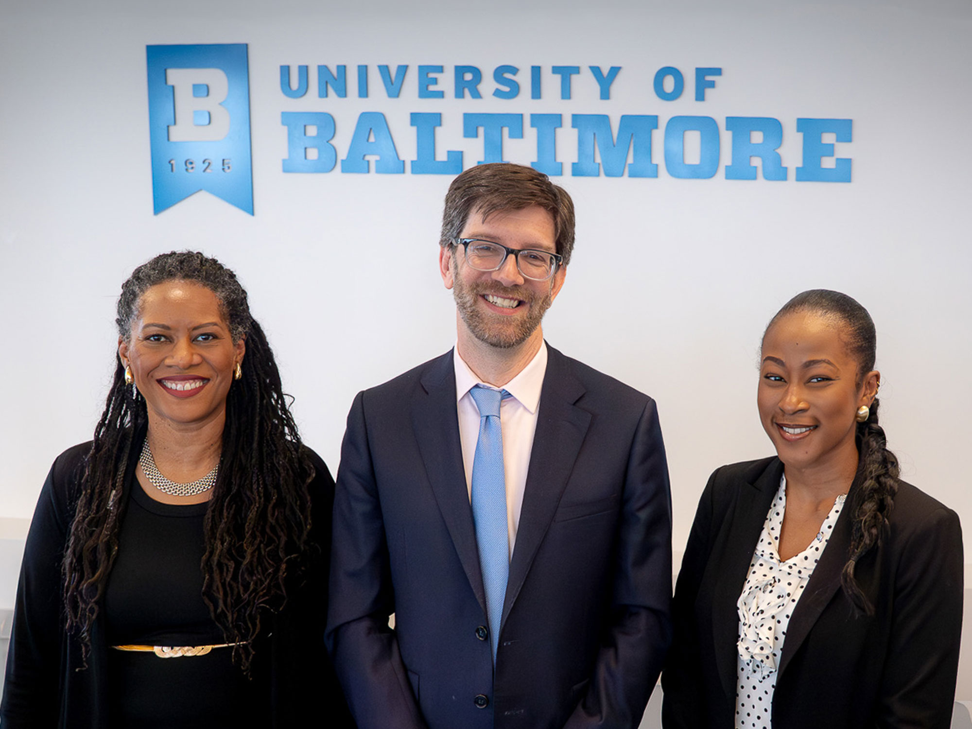 Three people stand together smiling in front of a University of Baltimore wall sign.