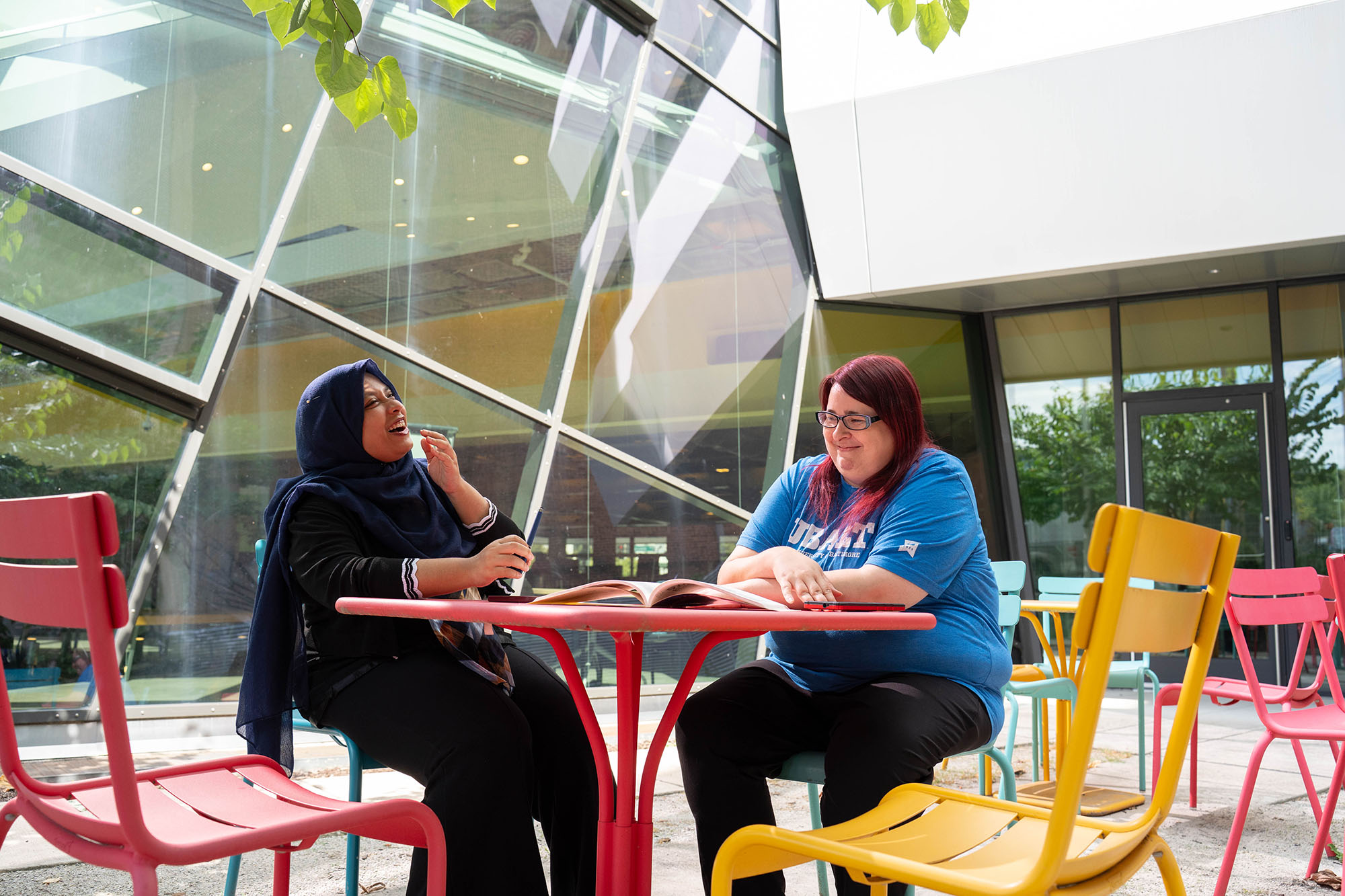 Two students sit outside the University of Baltimore library in an atrium, sharing stories and laughing.
