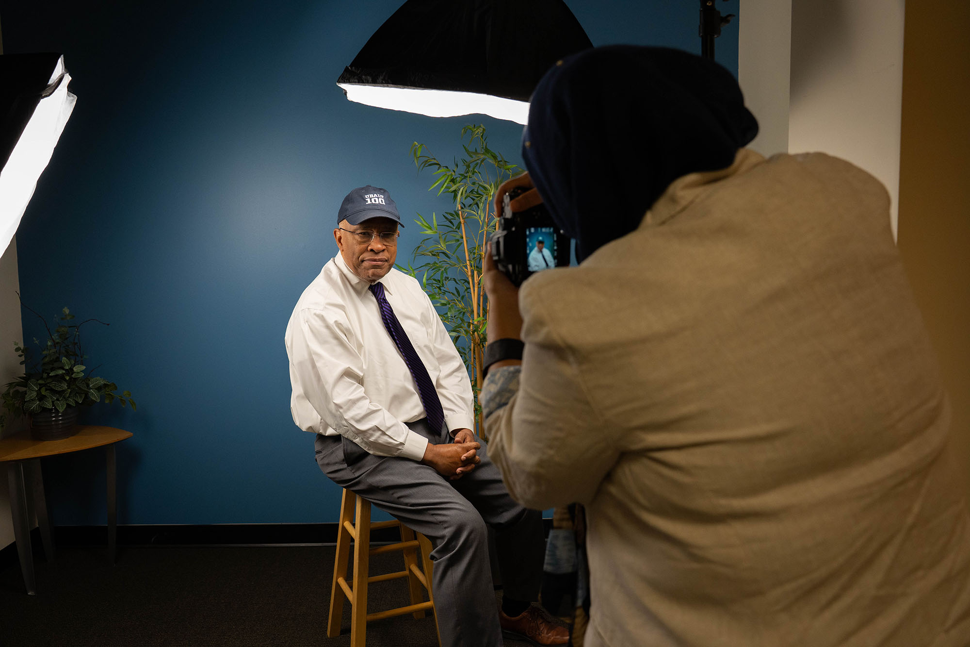 A student, back to camera, takes a picture of the UBalt president as he sits on a stool in a photo studio. 