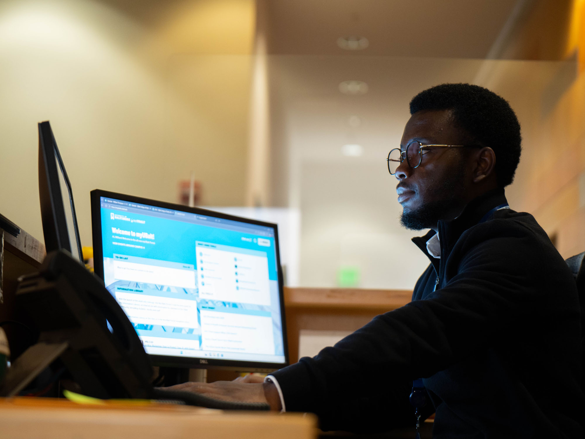 A student sits at an office front desk looking at one of two computer screens on the desk during his role as a student employee.