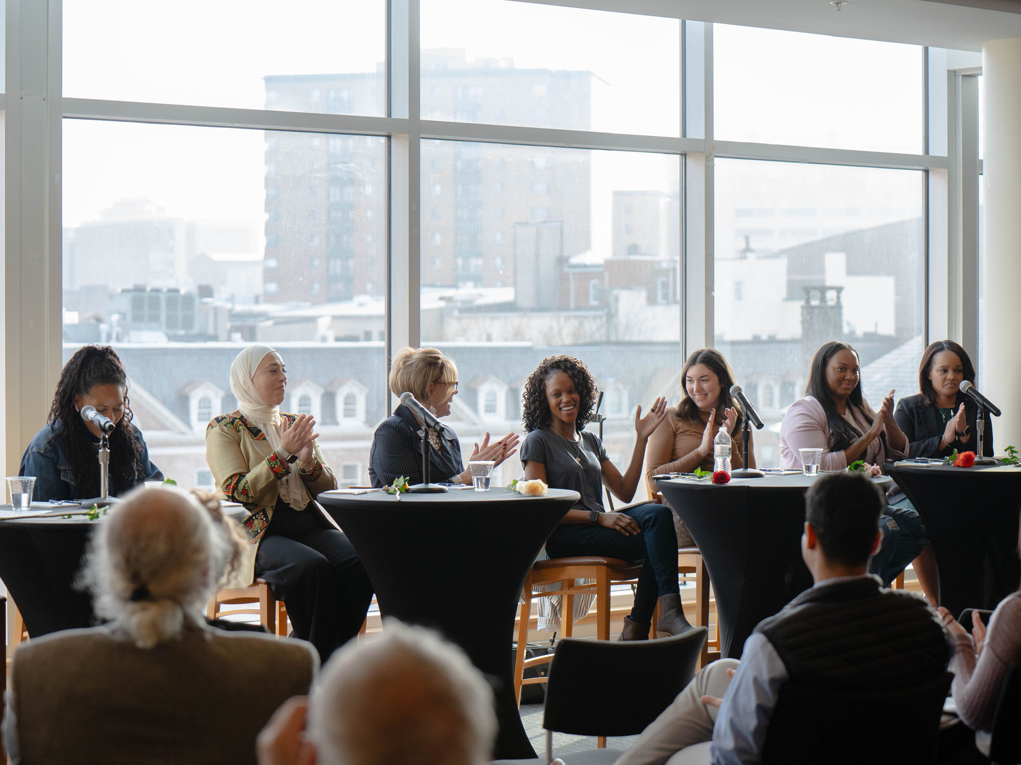 A group of women panelists applaud while one woman, at the center, raises her hand. An audience watches.