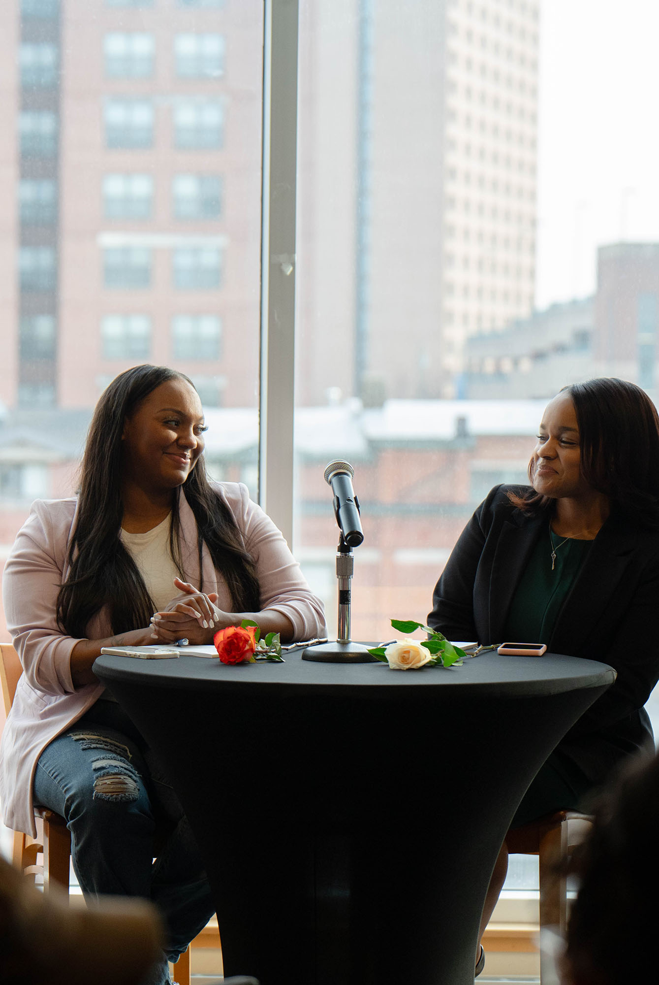 Two women on a panel, sitting at a table, share a smile.